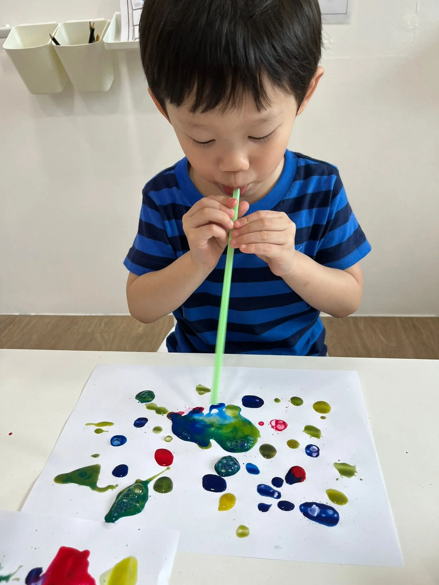 A young child in a blue striped shirt is blowing a green bubble wand into colorful paint splattered on a white paper.