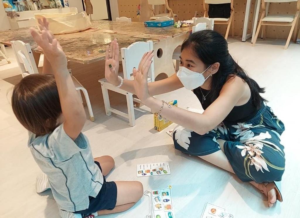 A woman wearing a face mask and a sleeveless dress is playing a game with a young boy in a room with wooden chairs and a table. They are giving each other high fives while sitting on the floor.