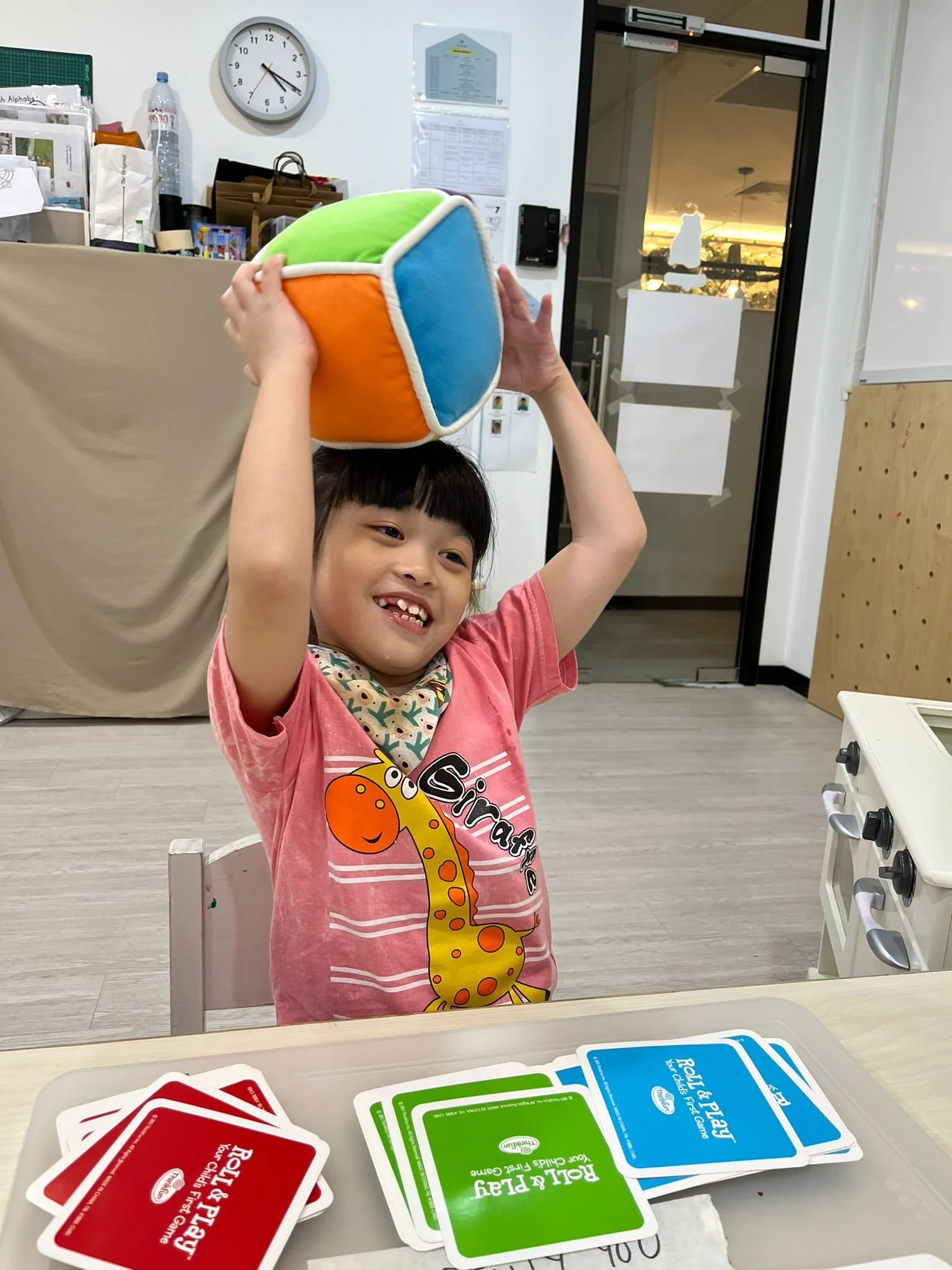A young girl with dark hair and a broad smile is holding a colorful plush cube above her head, playing a game of Uno in a room with a white wall, clock, and various objects in the background. There are Uno cards laid out on a table in front of her.