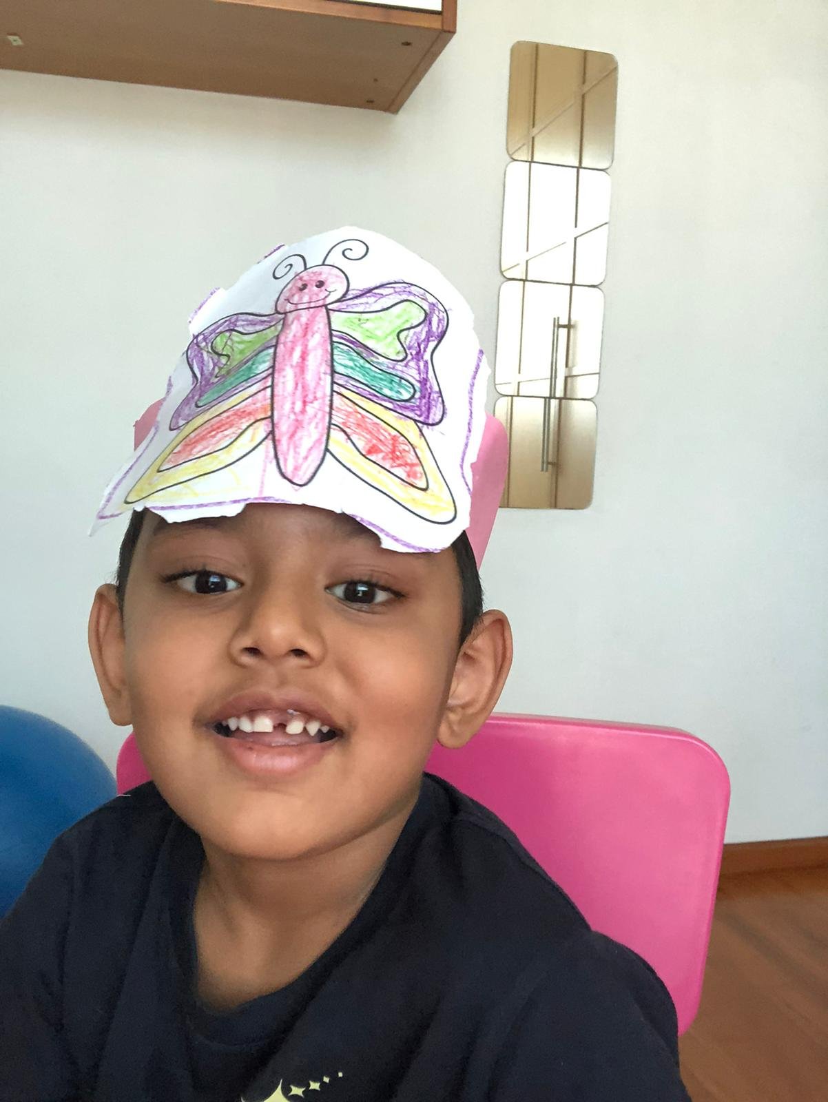A young boy with a missing front tooth, sitting in a pink chair, wearing a colorfully drawn paper butterfly hat on his head, smiling at the camera.