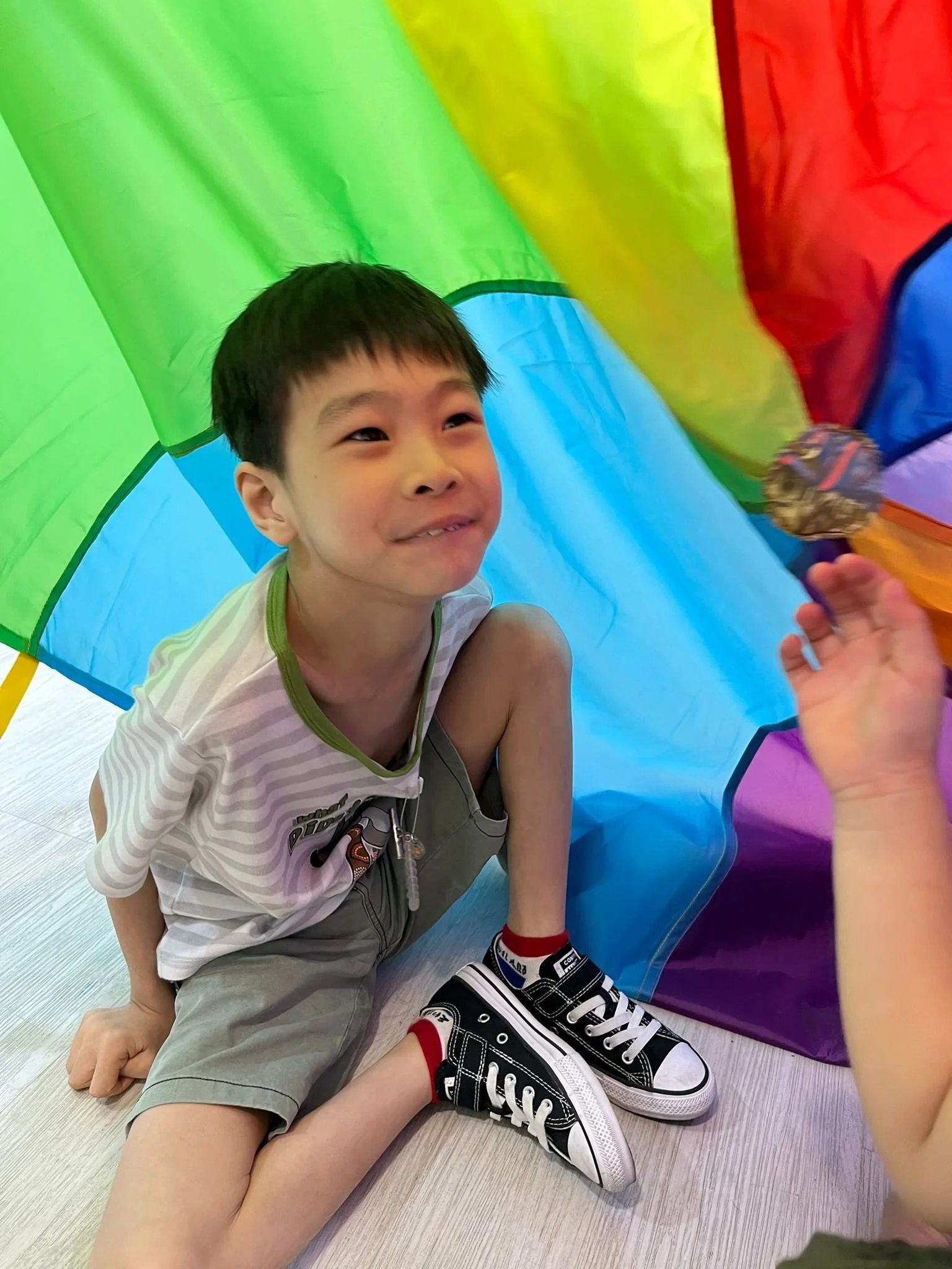 A young boy sitting on the floor inside a colorful, rainbow-colored play tunnel, smiling and holding a spoon in his hand. He is wearing a striped shirt, gray shorts, and black-and-white sneakers. Another person's hand is visible near the boy, holding the spoon.