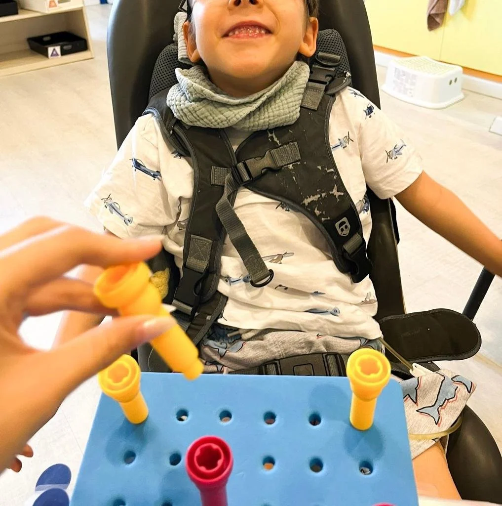 A young boy sitting in a wheelchair smiling, with an adult's hand holding colorful plastic game pieces over a blue game board in front of him.