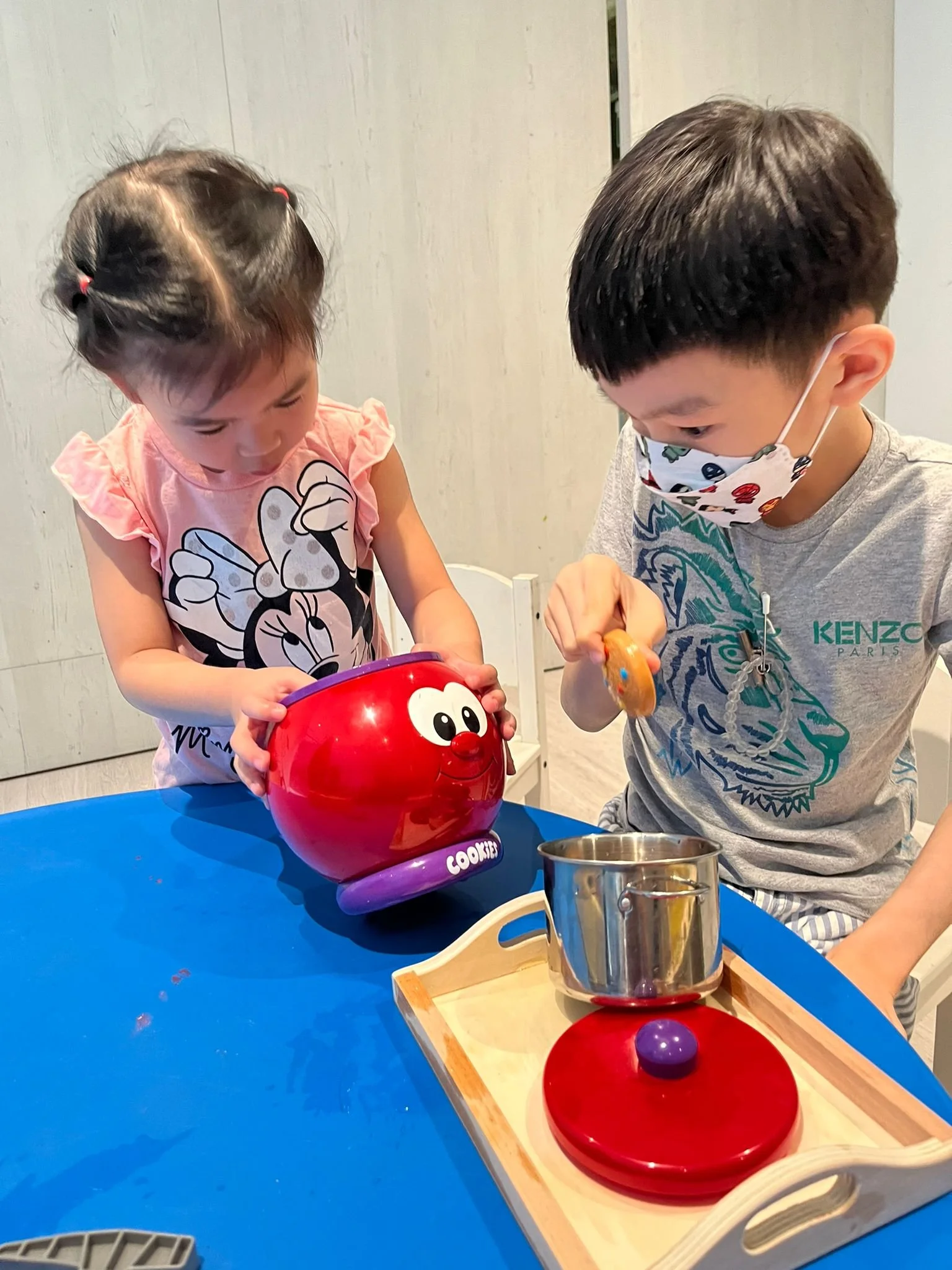 Two children, a girl and a boy, playing a cookie jar game at a blue table. The girl is holding a red container with a cartoon face, while the boy, wearing a face mask, is reaching into a wooden cookie jar game.