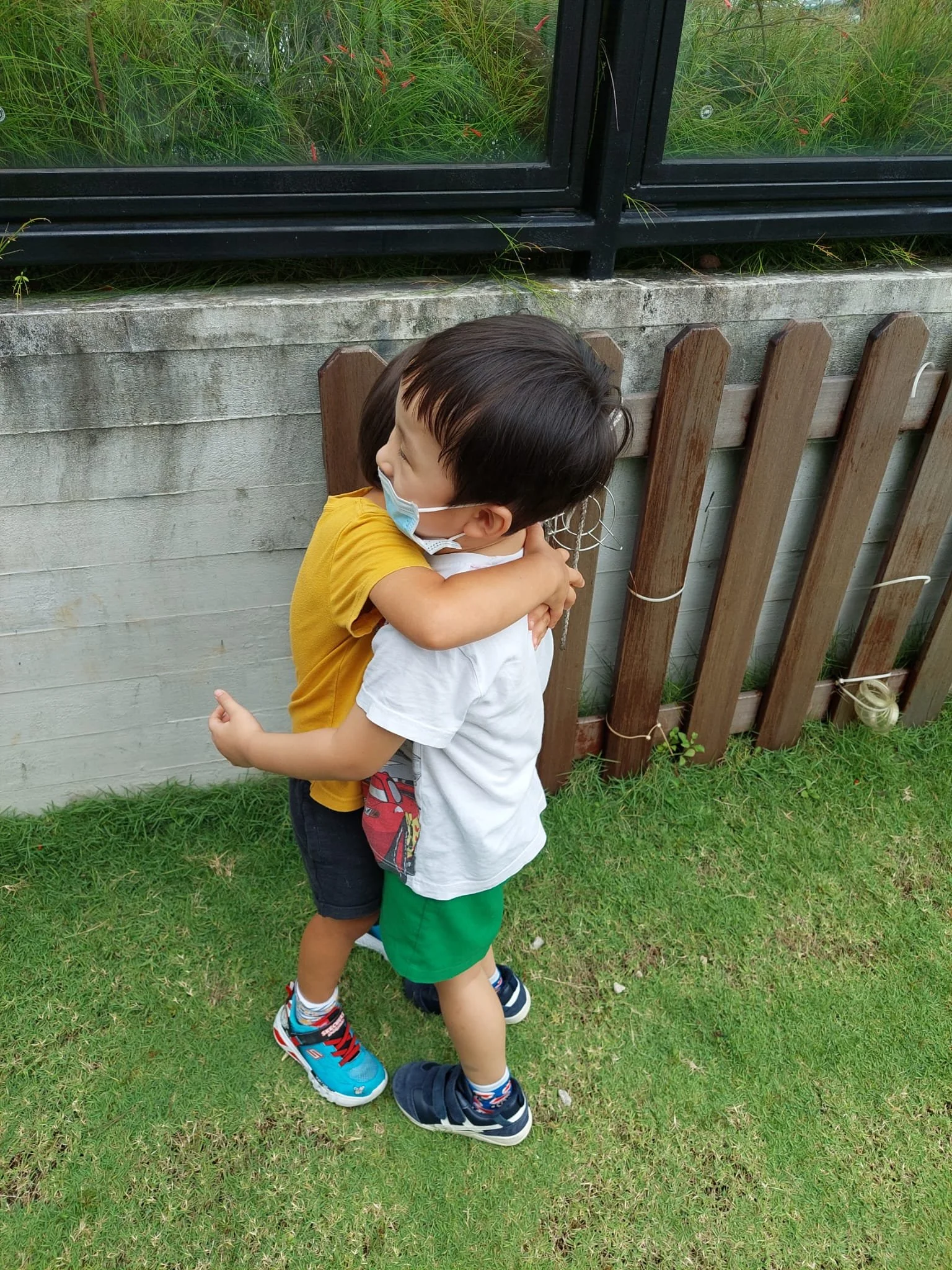 Two young boys hugging each other outdoors, wearing face masks, near a wooden fence and a window with green plants behind it.