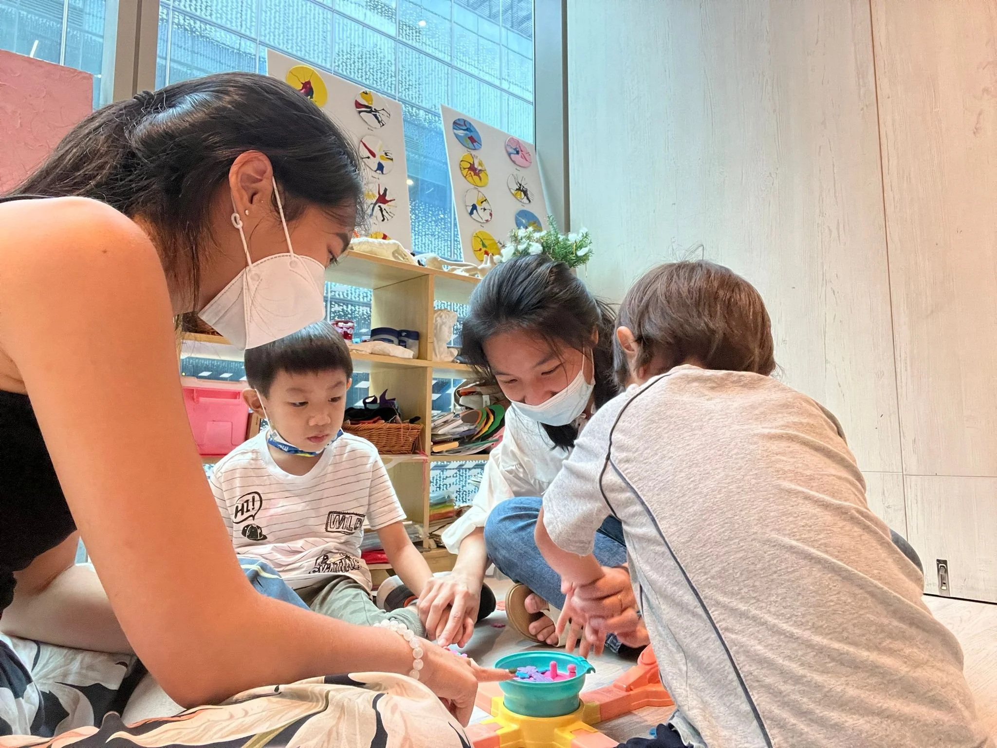 Group of people, including children and adults, engaged in a hands-on activity at a table in a room with large windows and wall art.