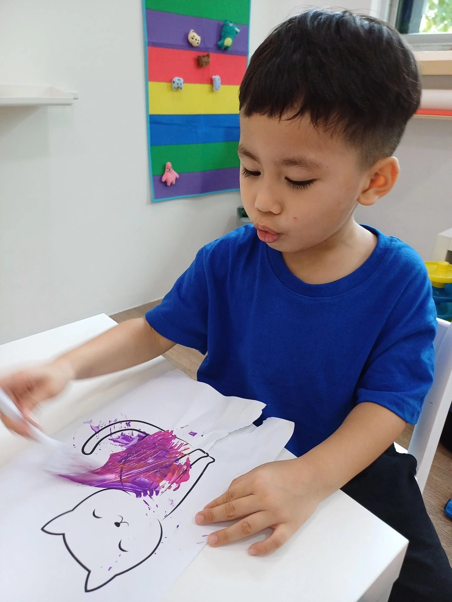 A young boy in a blue shirt is coloring on a paper with a unicorn drawing, using purple and pink paint. The boy is focused on his art, and in the background, there is a colorful bulletin board with small plush toys attached to it.