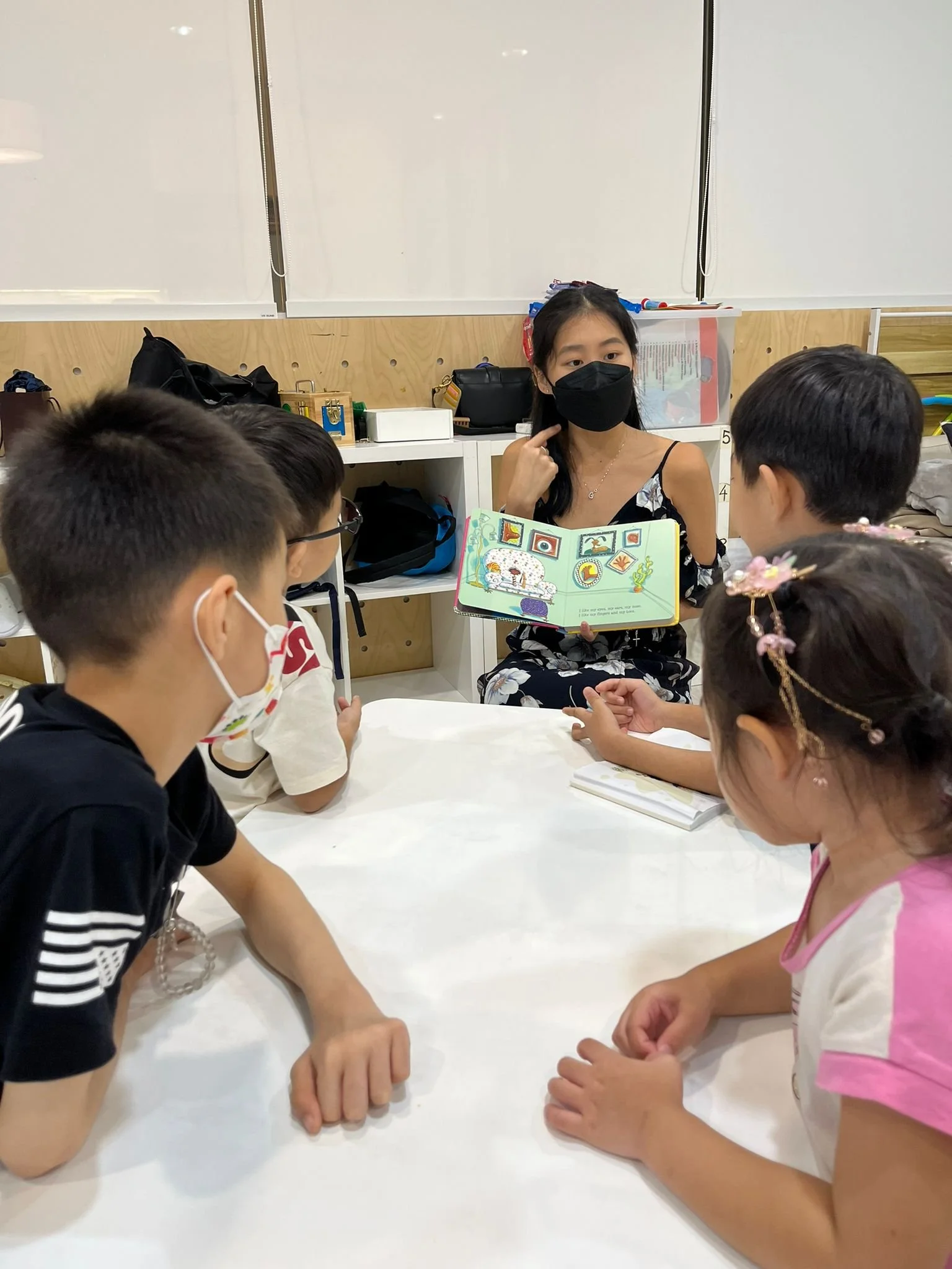 Children sitting around a table with a woman wearing a black face mask, holding a colorful children's book, in a classroom setting.