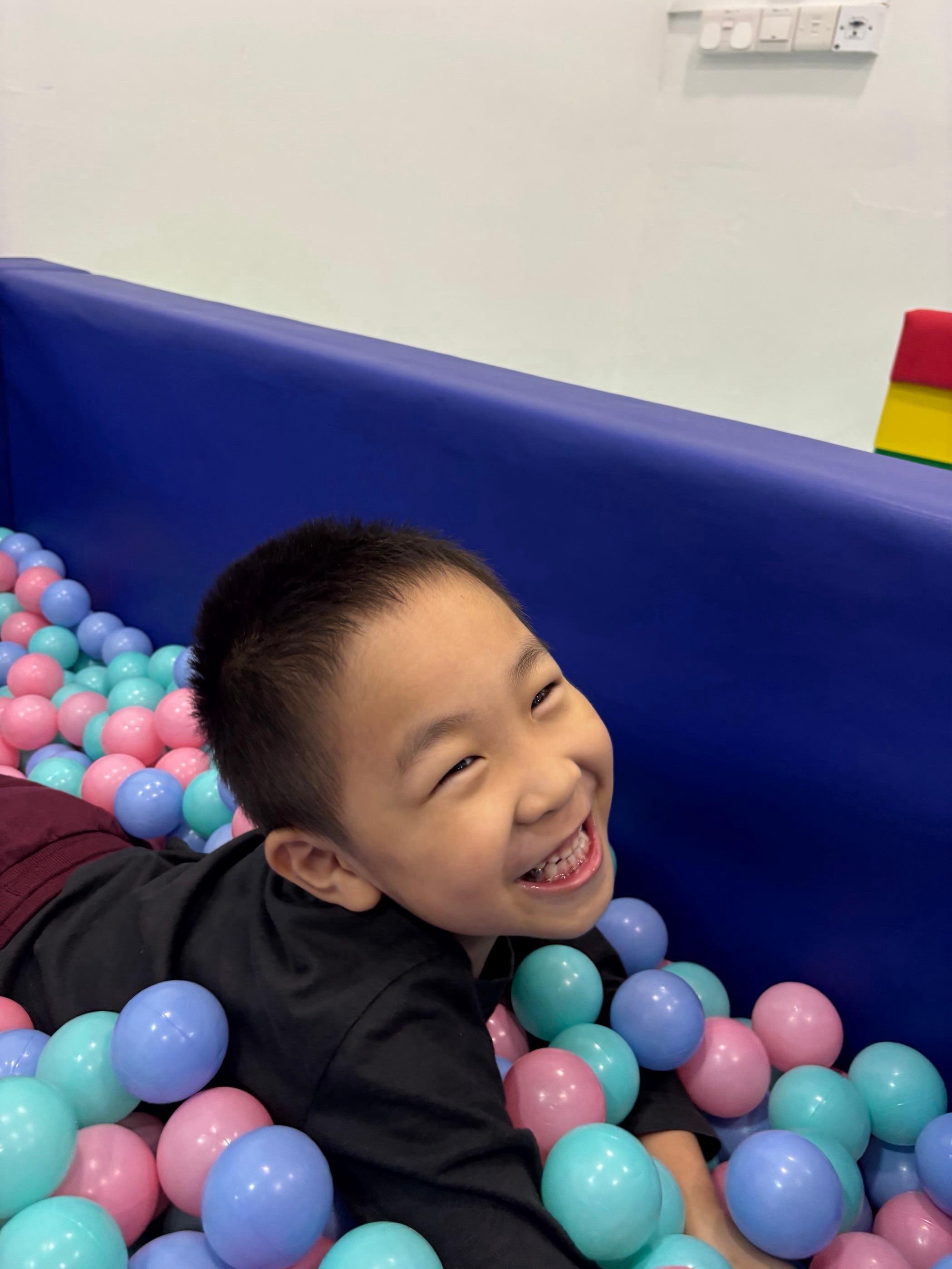 A young boy with short black hair smiling and laughing while sitting in a ball pit filled with pink, purple, and light blue plastic balls. The background includes a plain white wall with some electrical outlets and a colorful stacking block toy.