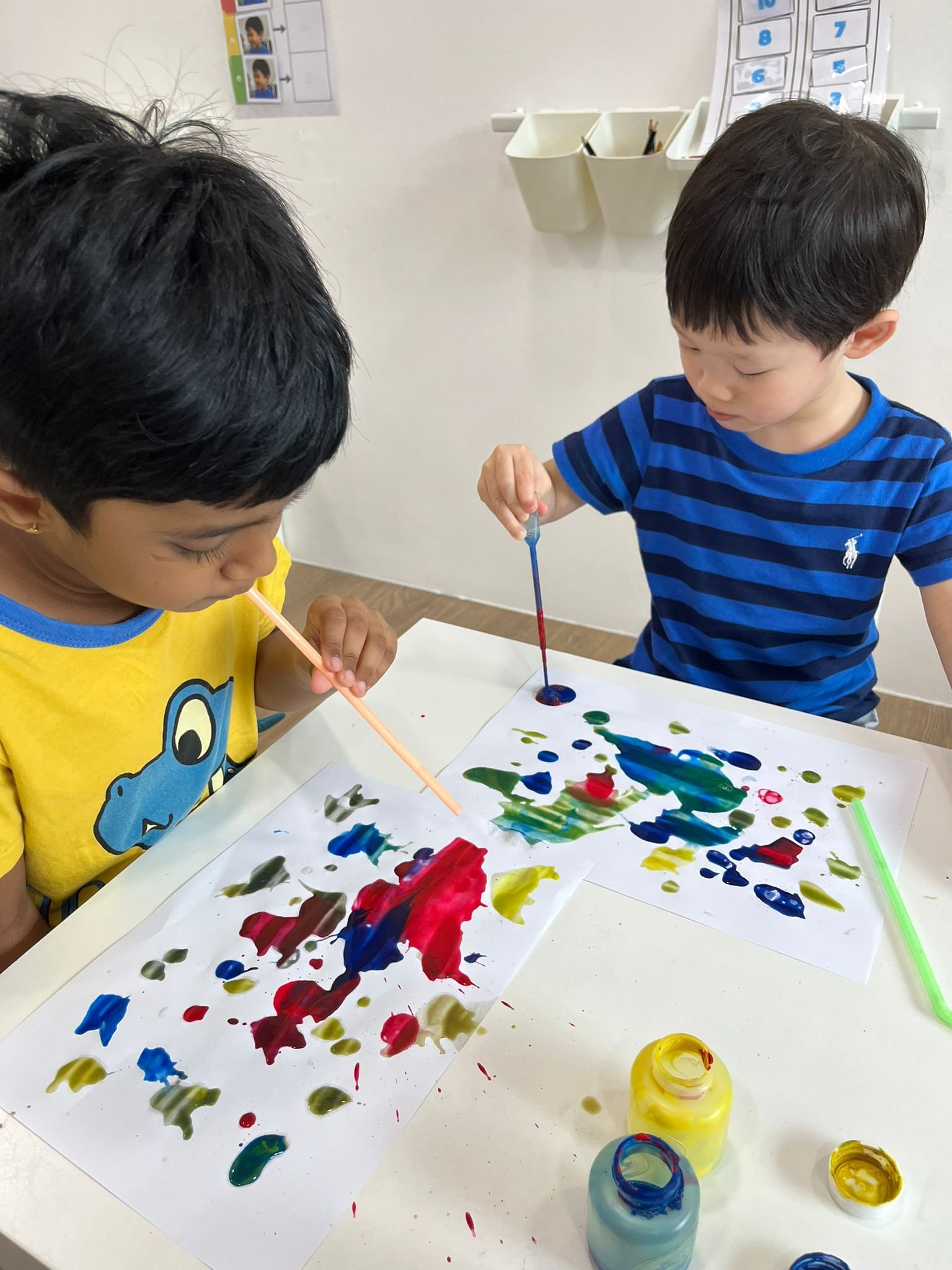 Two young children engaging in finger painting with colorful paints on large sheets of paper, with paint jars and straws on the table.