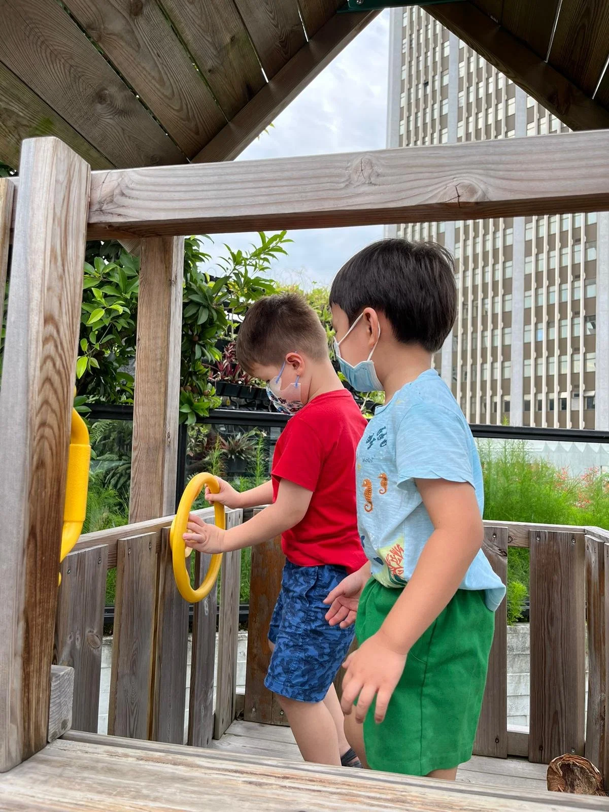 Two young boys wearing face masks are playing on a wooden outdoor structure. One boy in a red shirt and blue shorts holds a yellow ring, while the other boy in a light blue shirt and green shorts stands nearby. There are trees and a tall building in the background.