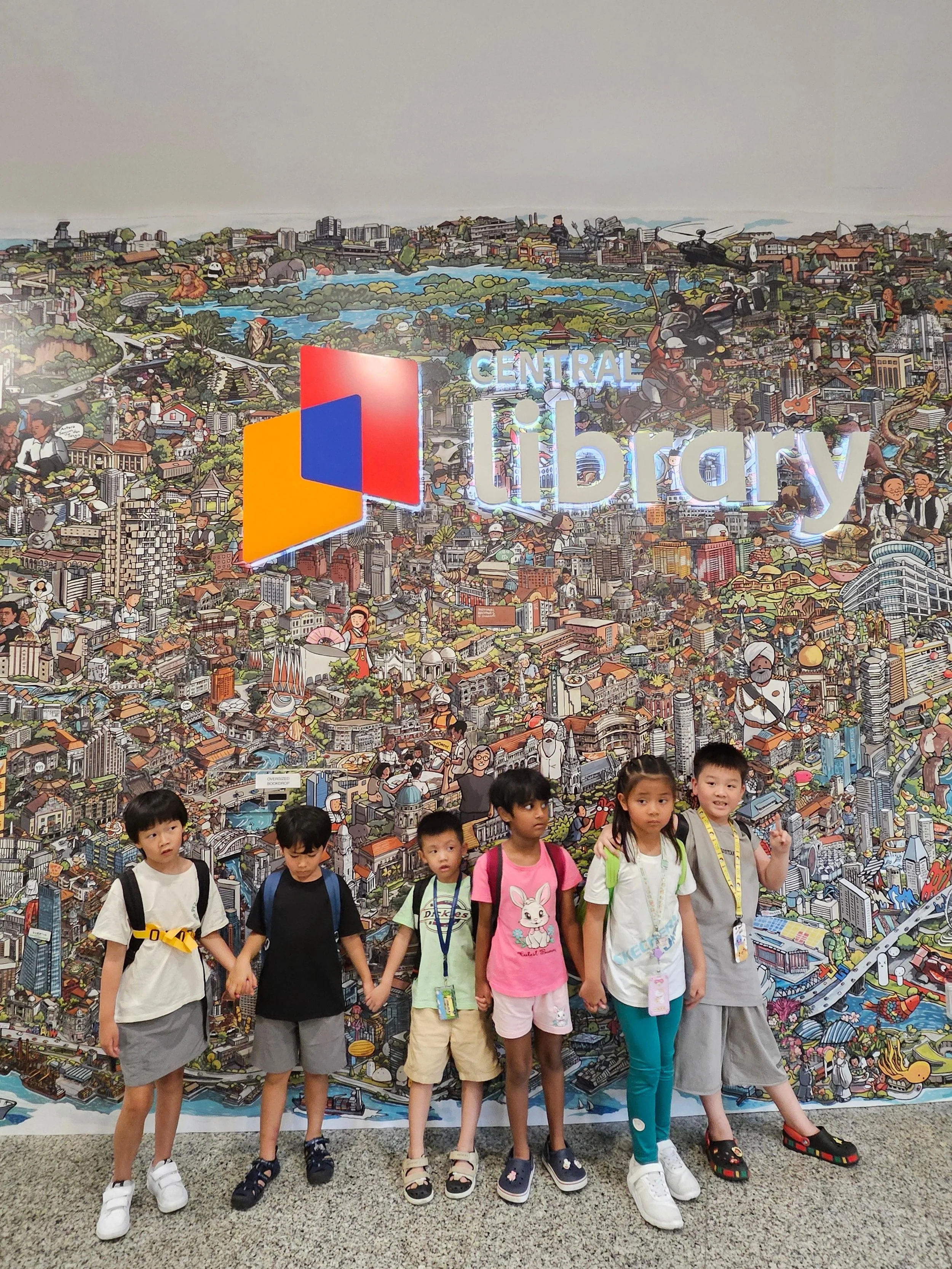 Group of children holding hands in front of a colorful cityscape mural at an indoor location.