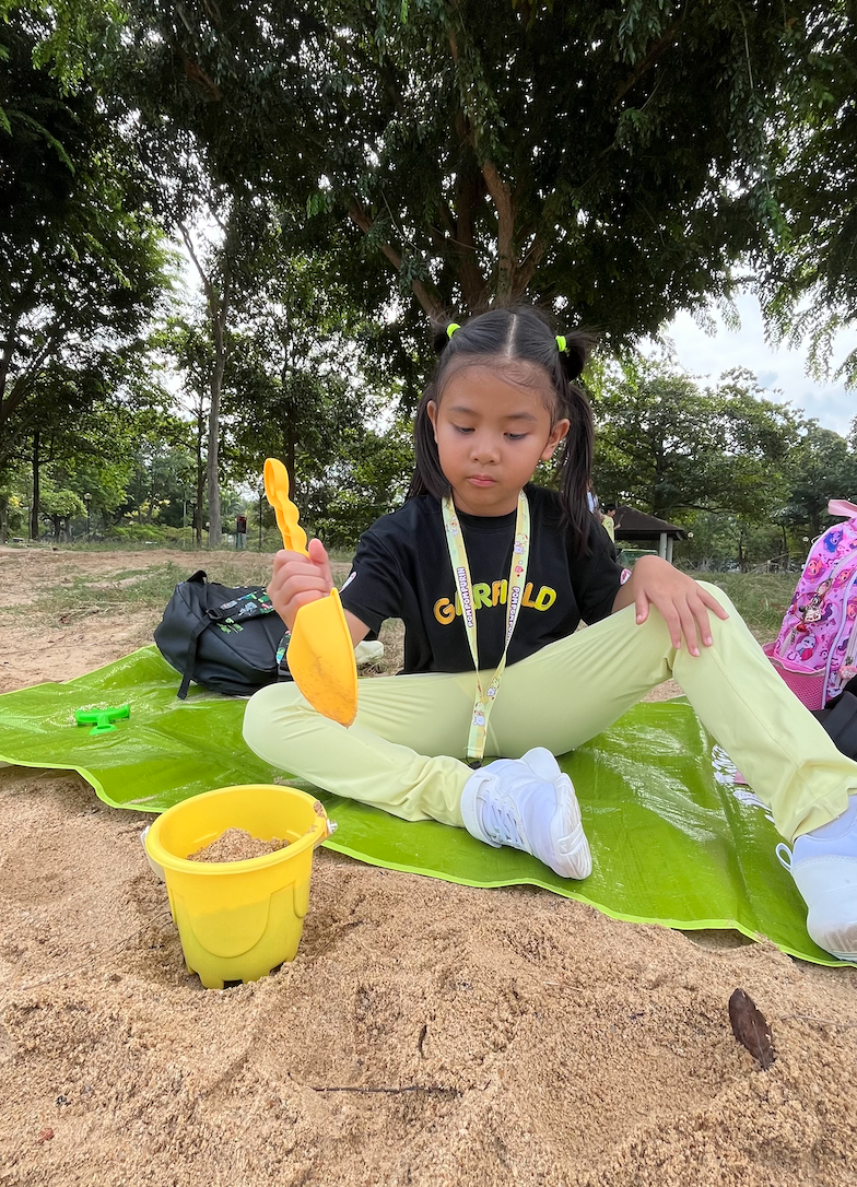 A young girl with pigtails sitting on a large green leaf on the ground, playing in a sandbox with a yellow toy shovel and a yellow bucket filled with sand. She is outdoors under a tree with other children and backpacks visible in the background.