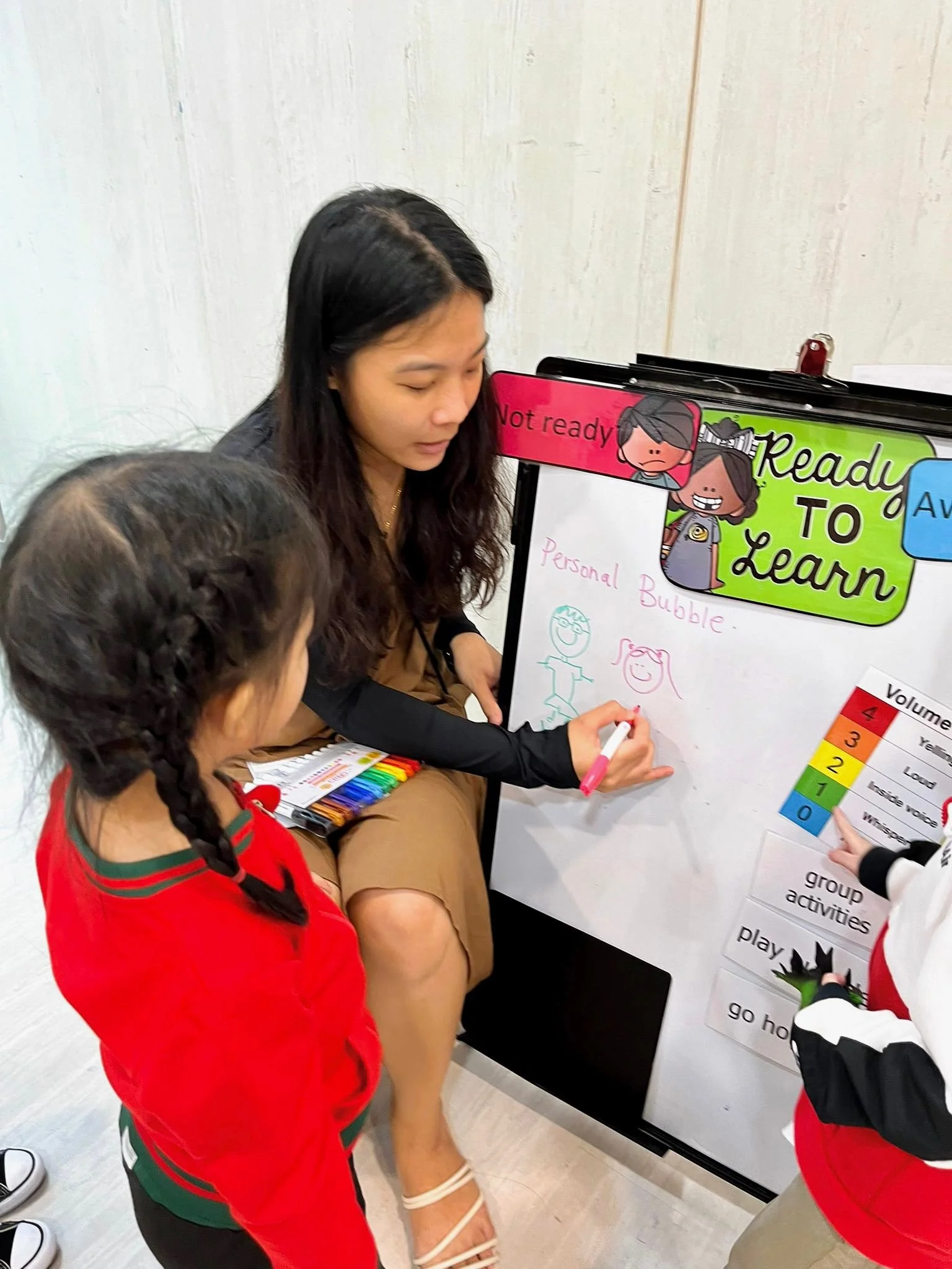 A woman sitting on a bench engaging with children in a learning activity involving a whiteboard, markers, and educational posters, with some children standing nearby.