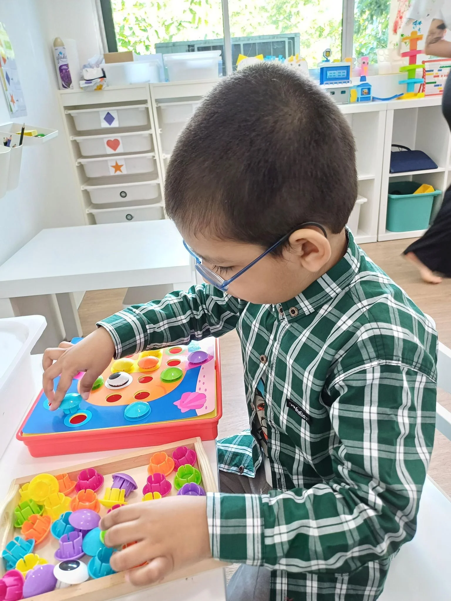 A young boy playing a board game with colorful plastic pieces in a room with shelves and toys in the background.