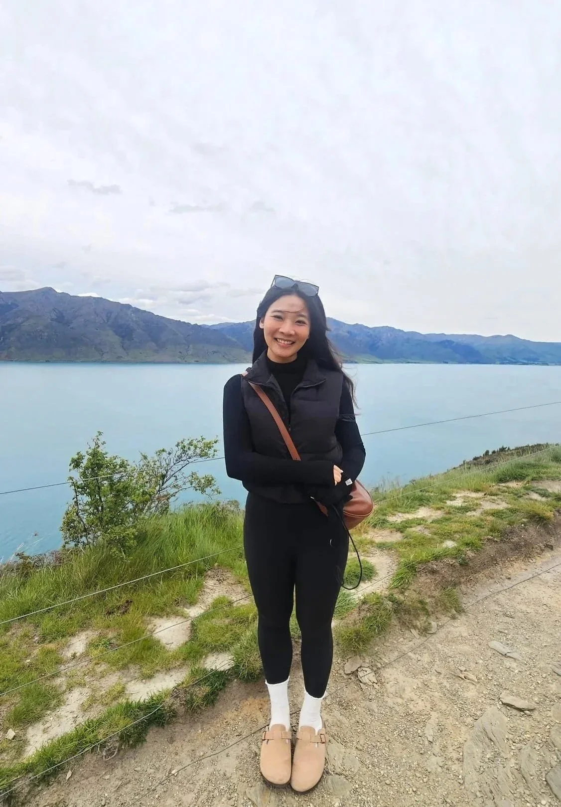 A woman standing outdoors near a large body of water with mountains in the background, smiling at the camera.