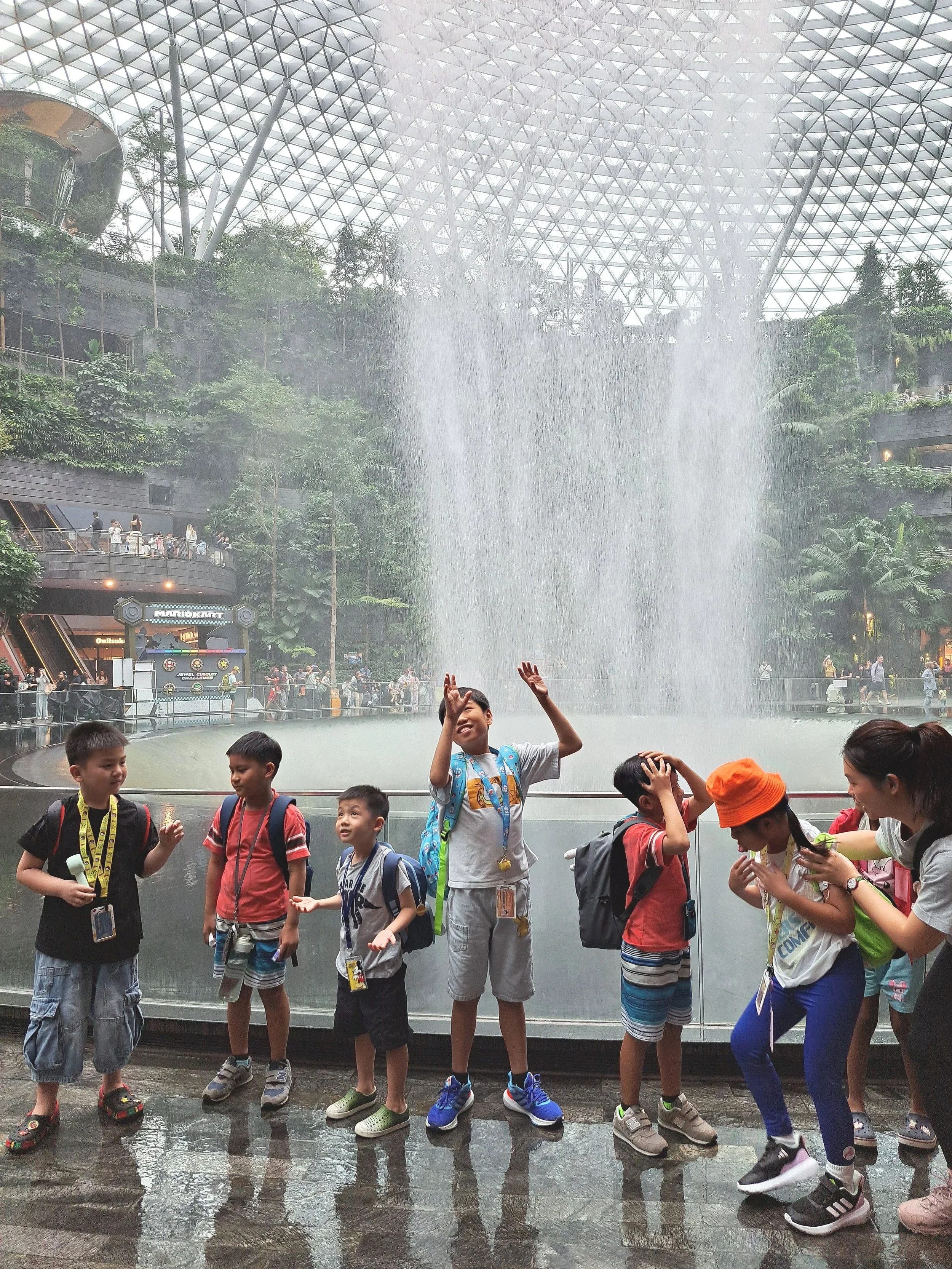 A group of children and a woman standing in front of a large indoor water fountain with water cascading down behind them. The indoor space has a glass-domed ceiling and lush greenery.