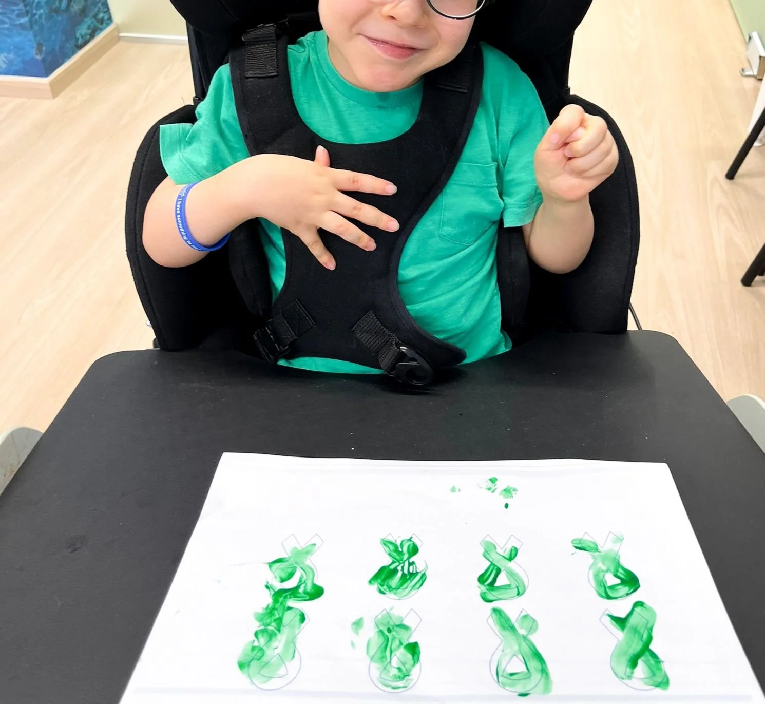 Child sitting at a table with green paint on paper, creating painted dollar signs, wearing a bright green shirt, glasses, and a black harness.