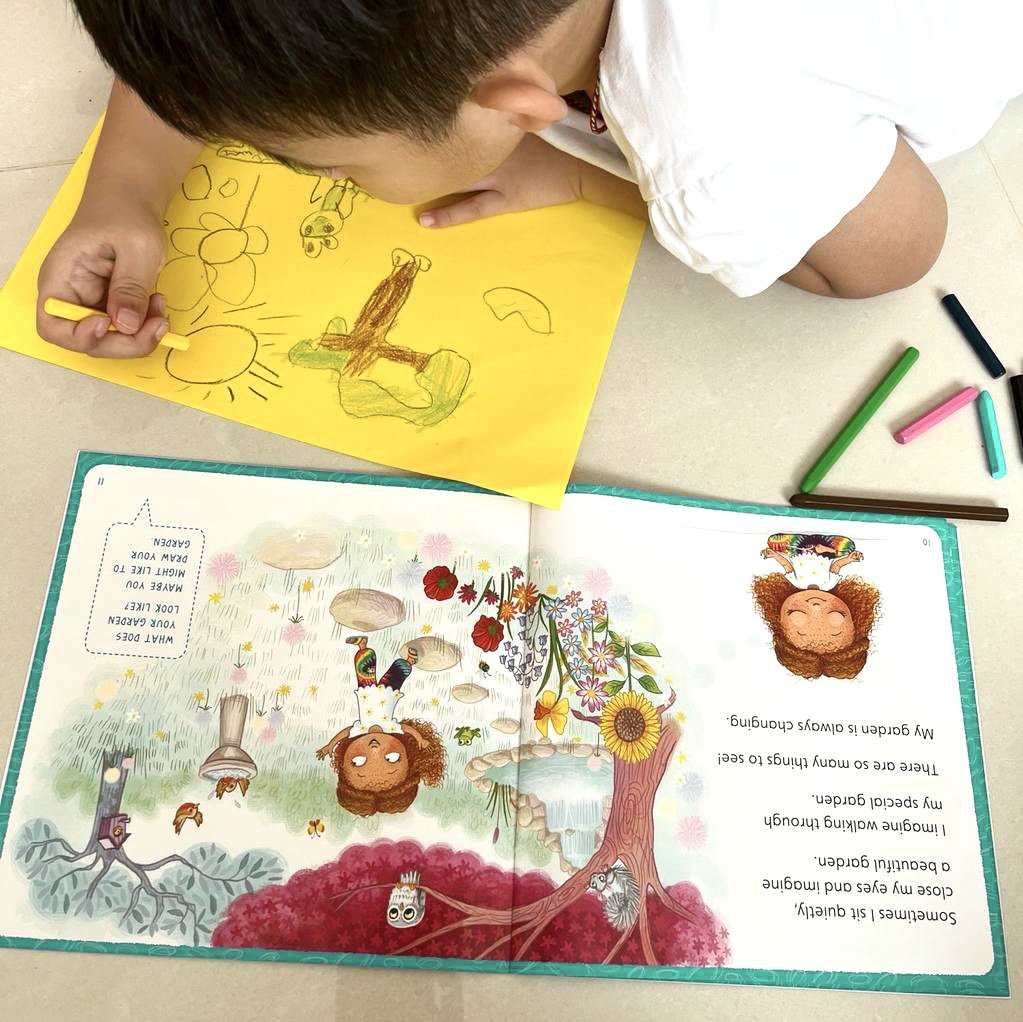 A child drawing a picture on yellow paper with colored markers, and a colorful illustrated children's book open on a table showing a girl in a rainbow dress standing in a garden.