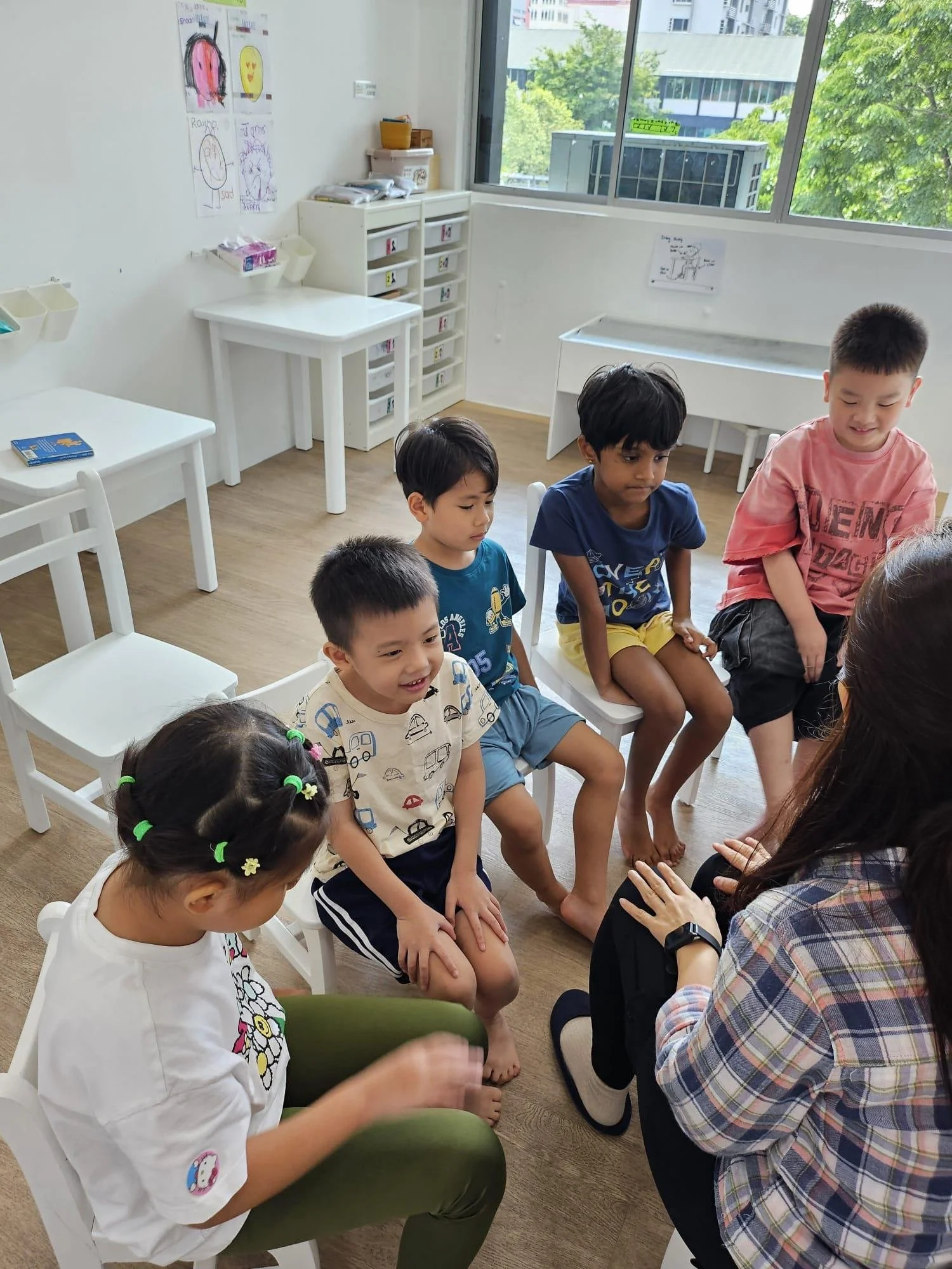 A group of children sitting on chairs in a classroom, with a woman engaging with them. The classroom has white walls, children's drawings on the wall, and large windows showing trees and buildings outside.