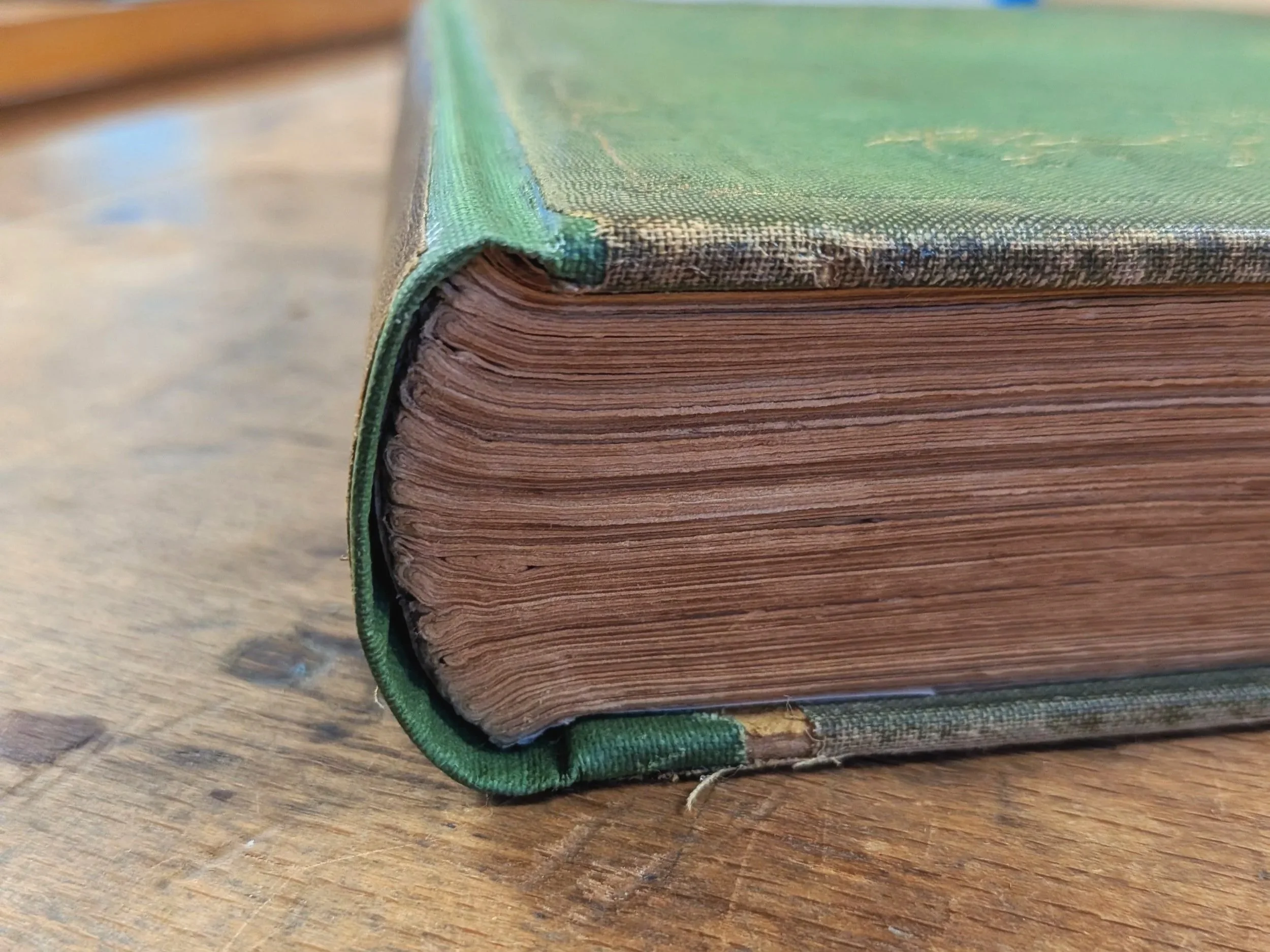 Close-up of an old, thick, leather-bound book with a green fabric cover and visible stacked pages resting on a wooden surface. This is an after picture showing a completed repair of the spine material.