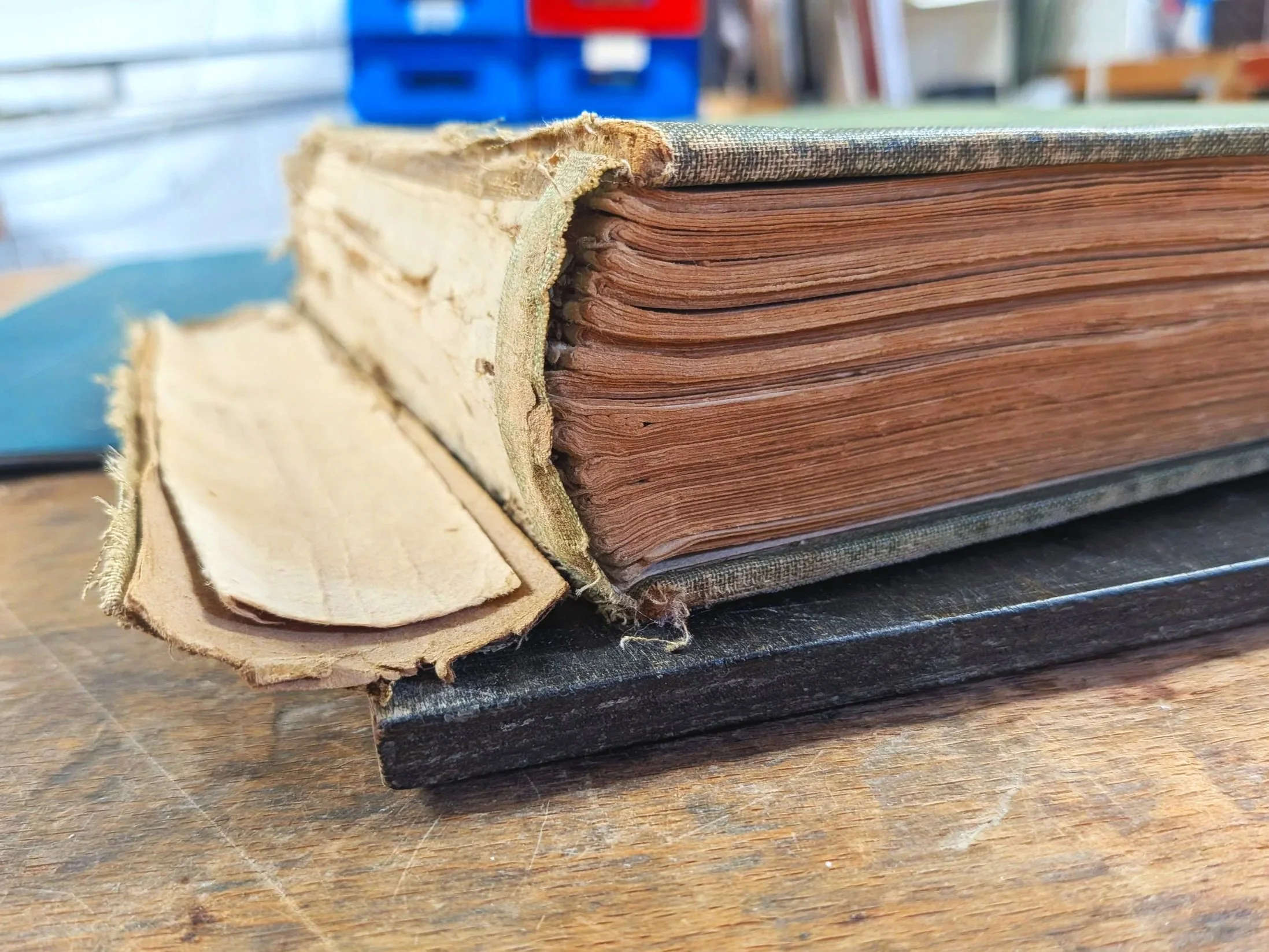 Old, worn book with frayed edges and yellowed pages resting on a wooden surface. This is a before picture, showing the damage to the spine and cover boards.