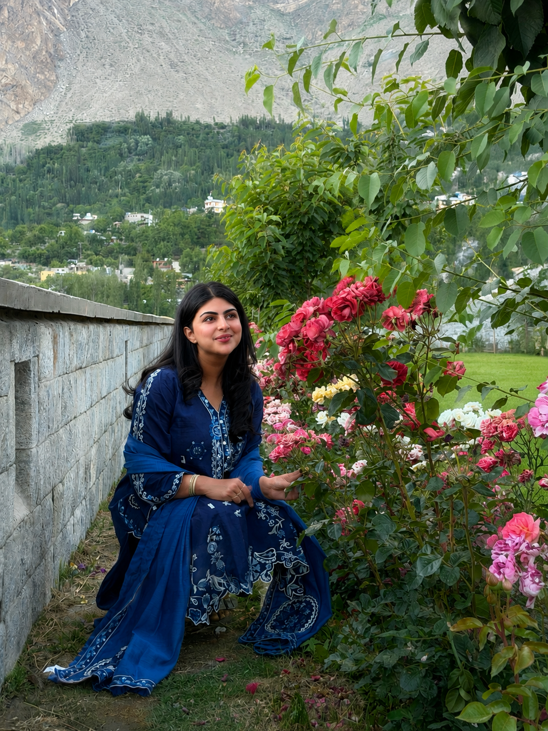 A woman in a blue traditional dress squatting near colorful flowering bushes in a garden with a mountainous landscape in the background.