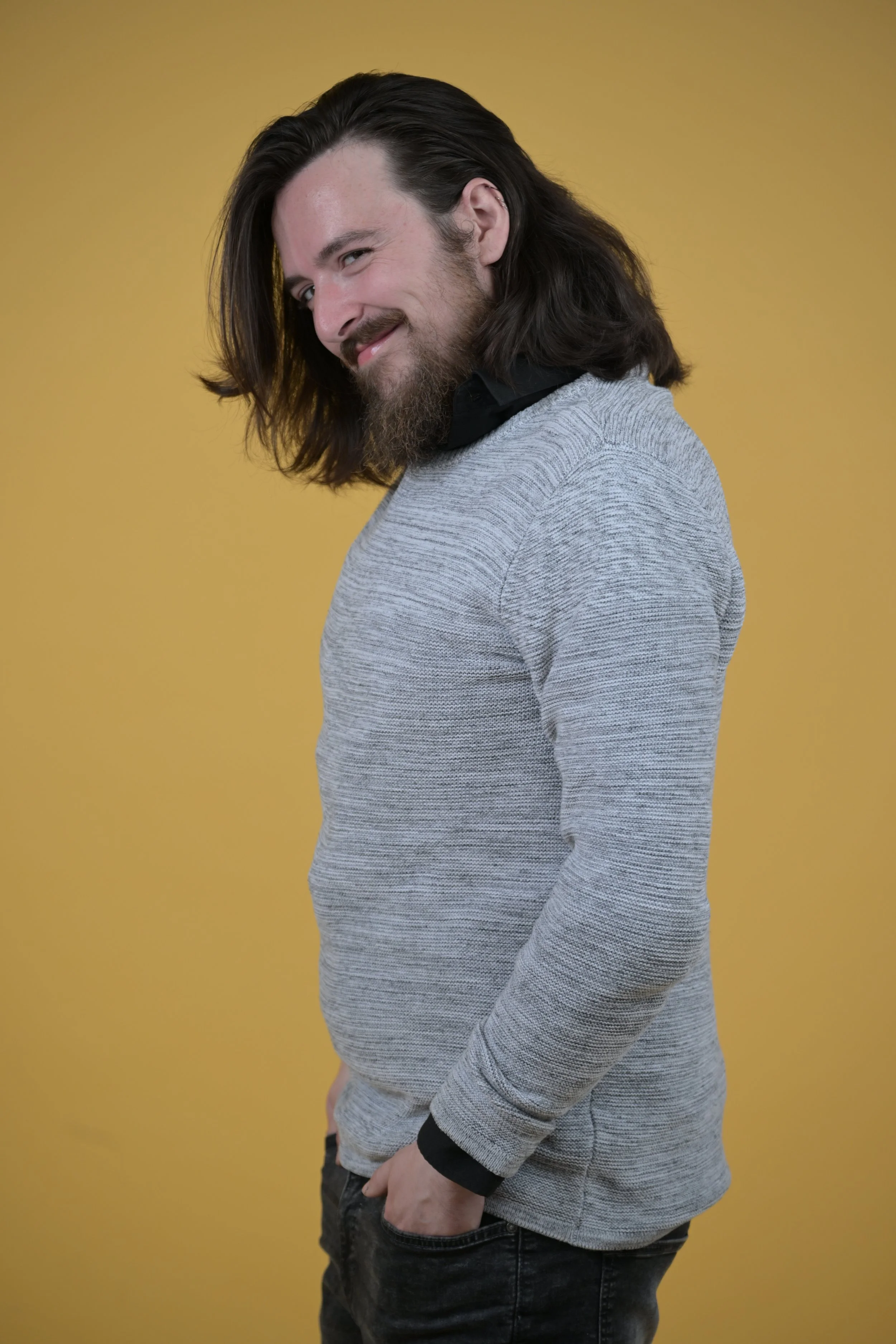 A young man with long dark hair, a beard, and a mischievous smile, standing against a yellow background, wearing a gray sweater over a black shirt.