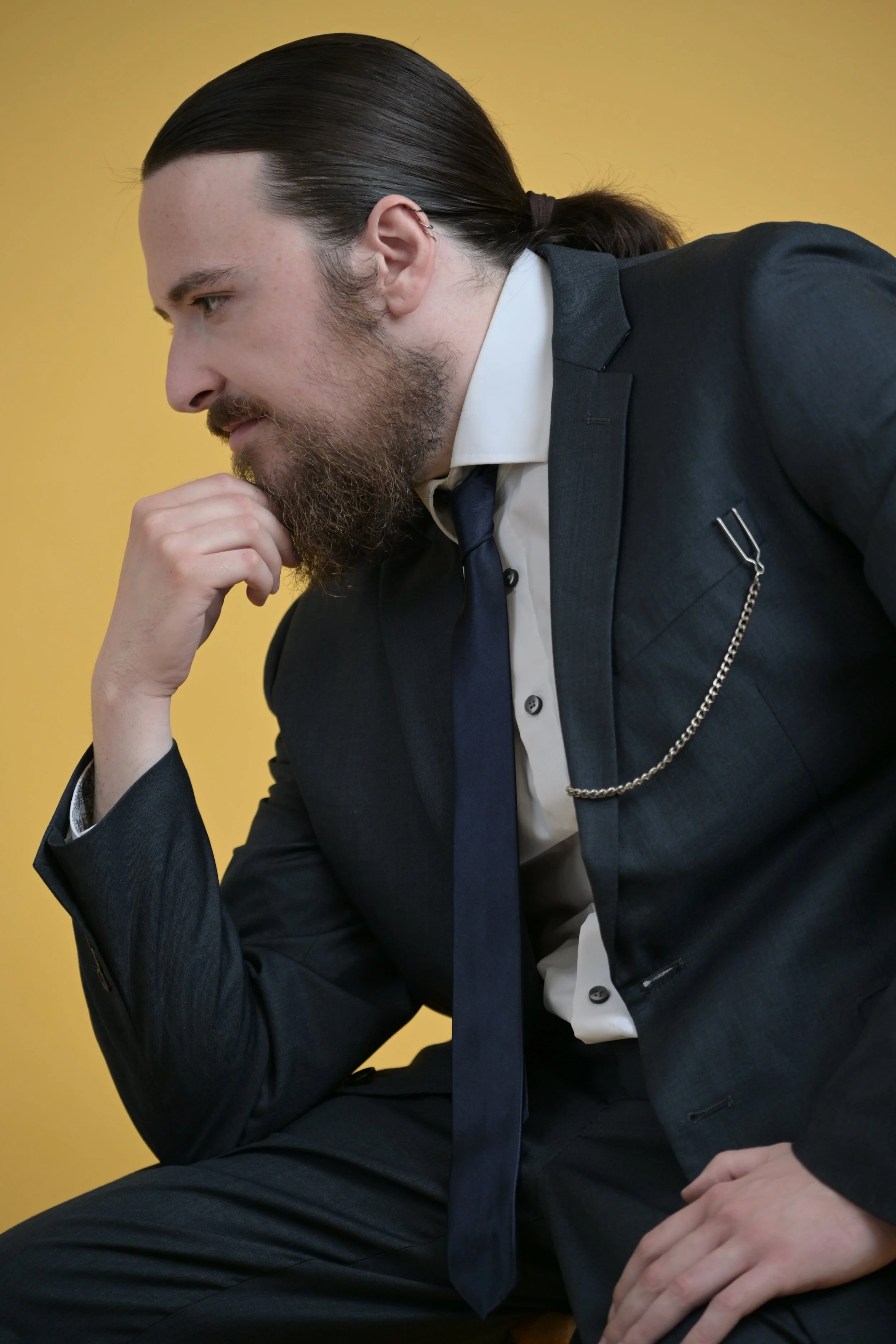 A man dressed in a dark suit with a chain accessory, white shirt, and navy tie, sitting against a yellow background. He has long dark hair tied back and a full beard, resting his chin on his hand and looking contemplative.