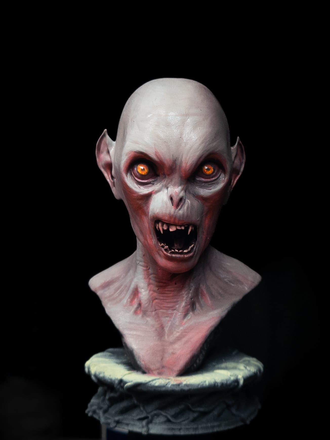 A sculpture or mask of a terrifying vampire or monster with pale flesh, bald head, pointed ears, glowing orange eyes, sharp fangs, and an angry expression, displayed on a stand against a black background.