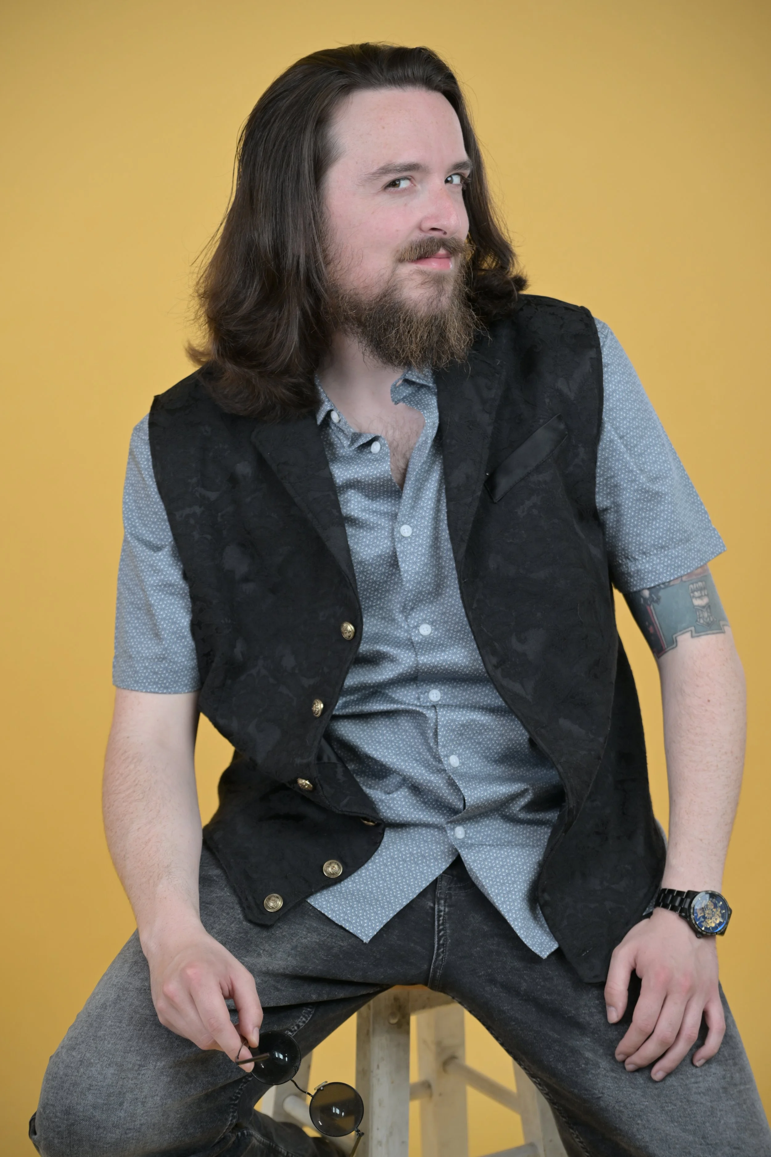A man with long brown hair, beard, and tattoo on his left arm, sitting on a wooden stool against a yellow background, wearing a gray shirt, black vest with gold buttons, and holding sunglasses in his right hand.