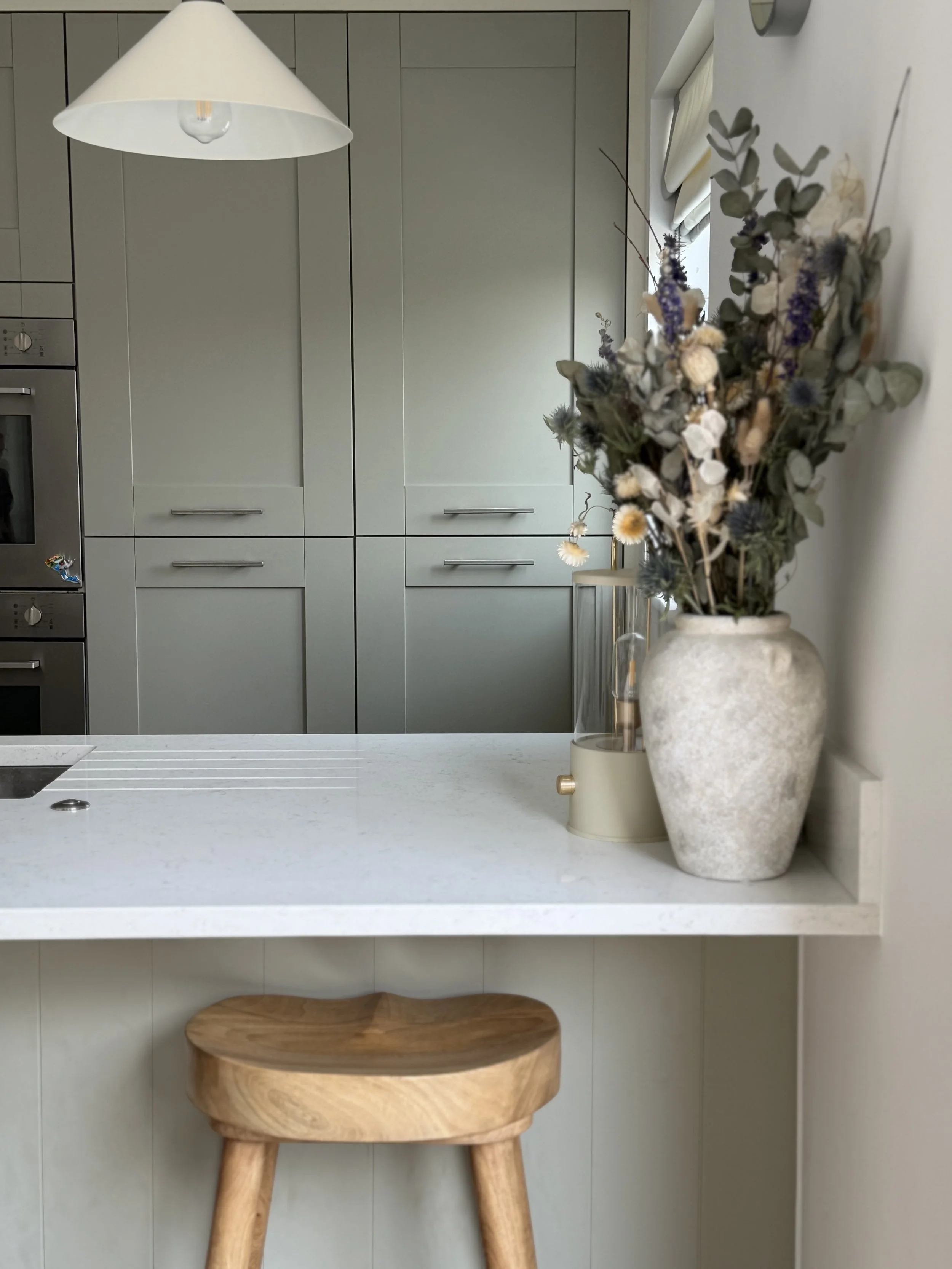 A kitchen scene featuring a white countertop with a large beige vase holding dried flowers, a small beige candle holder, and a wooden stool with rounded edges. In the background, there are gray cabinets, a window with curtains, and a white wall with a thermostat.