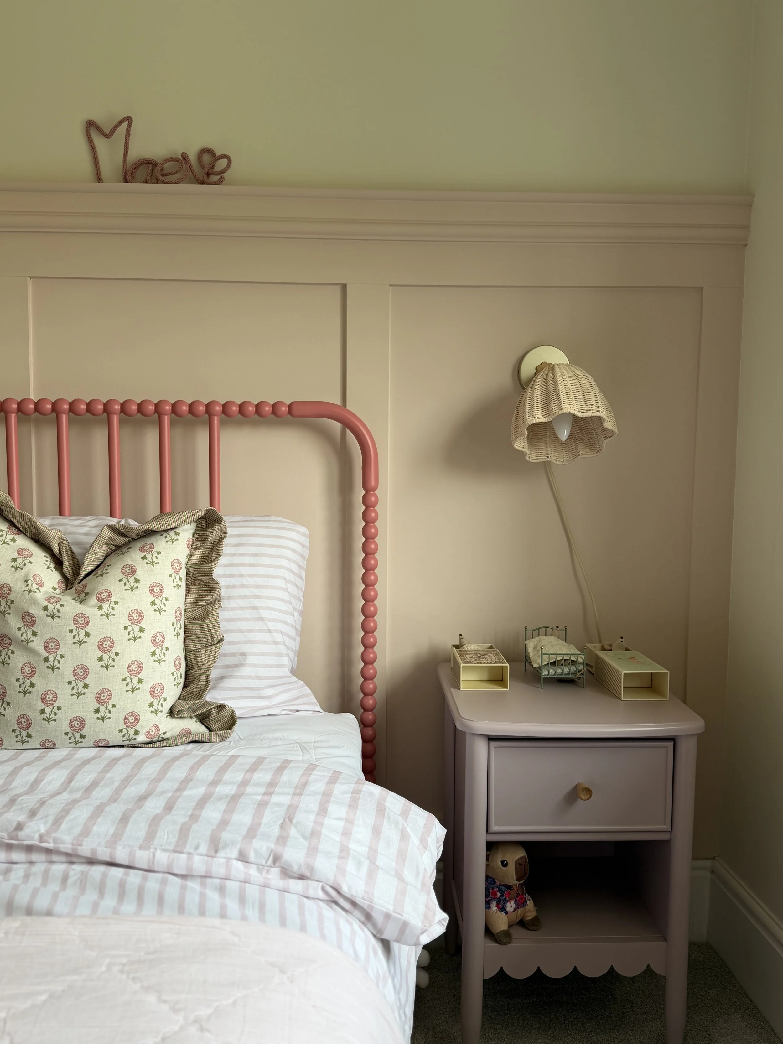 A bedroom corner featuring a bed with white and pink striped sheets, a pink bed frame, a floral pillow, a beige nightstand with a drawer, small decorative items, and a wall-mounted lamp with a wicker shade. There is a decorative word 'Love' on the green wall above the bed.