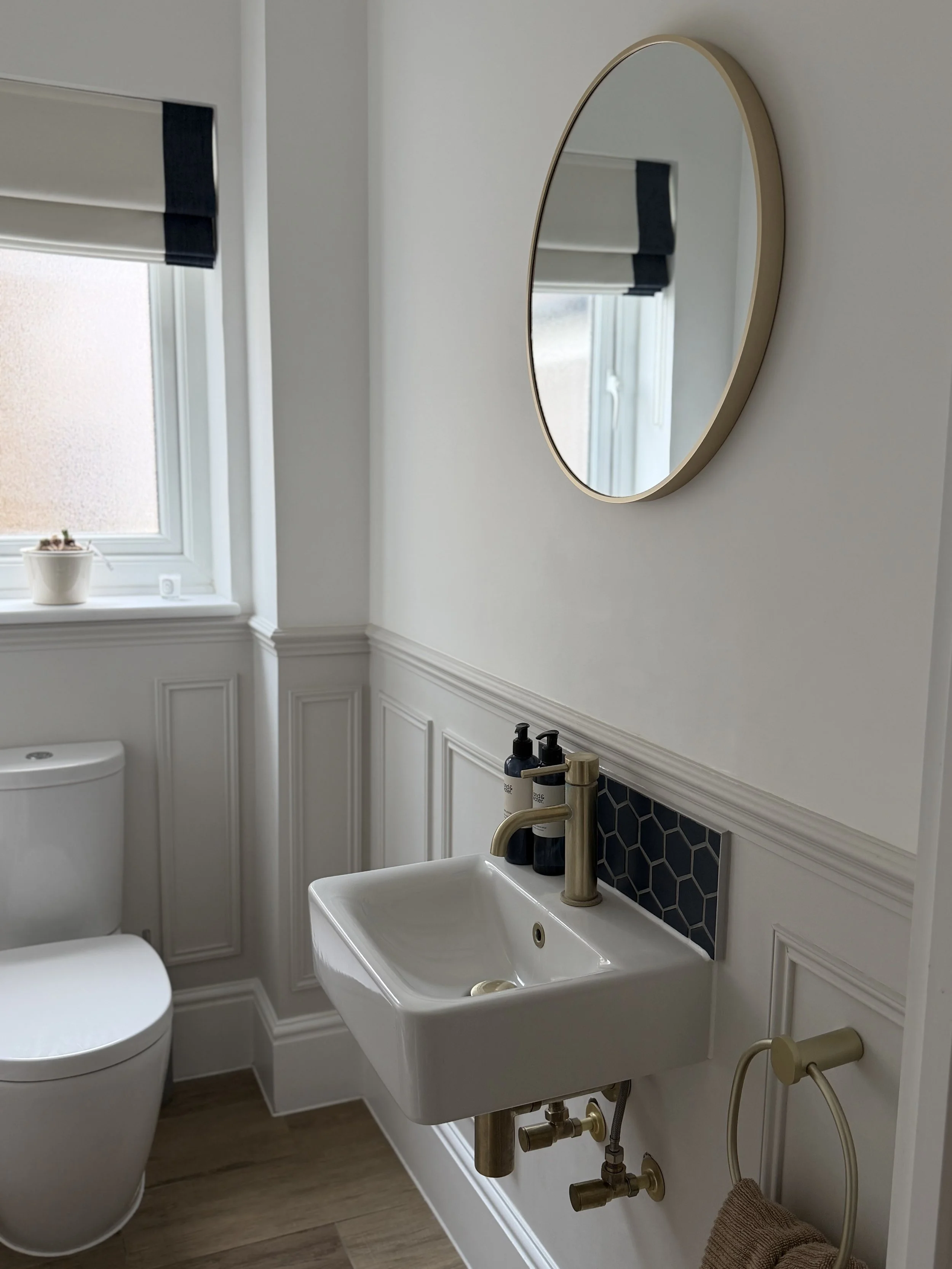A small bathroom with a white wall-mounted sink, a round mirror, and a toilet, with a window above the toilet and a towel hanging on a ring under the sink.