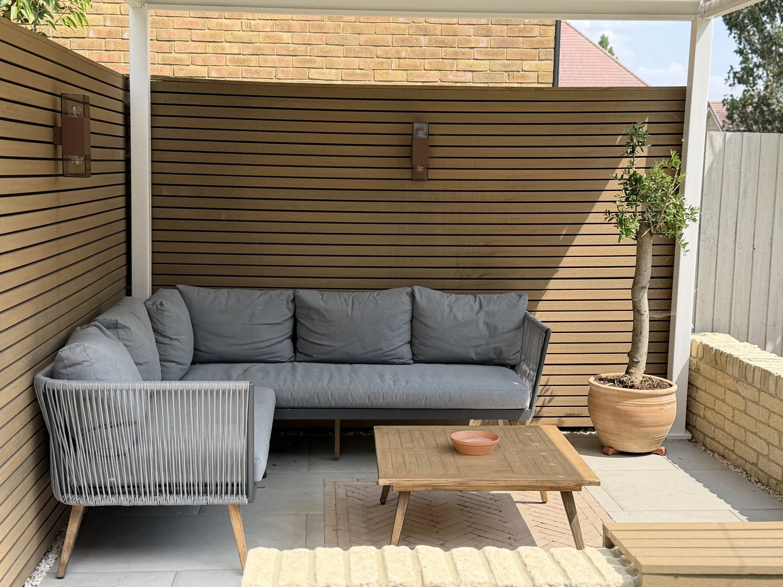 An outdoor patio area with a wooden privacy fence and a sofa with gray cushions. There is a small wooden table with a pink bowl on it, a potted tree, and a smaller gray armchair.