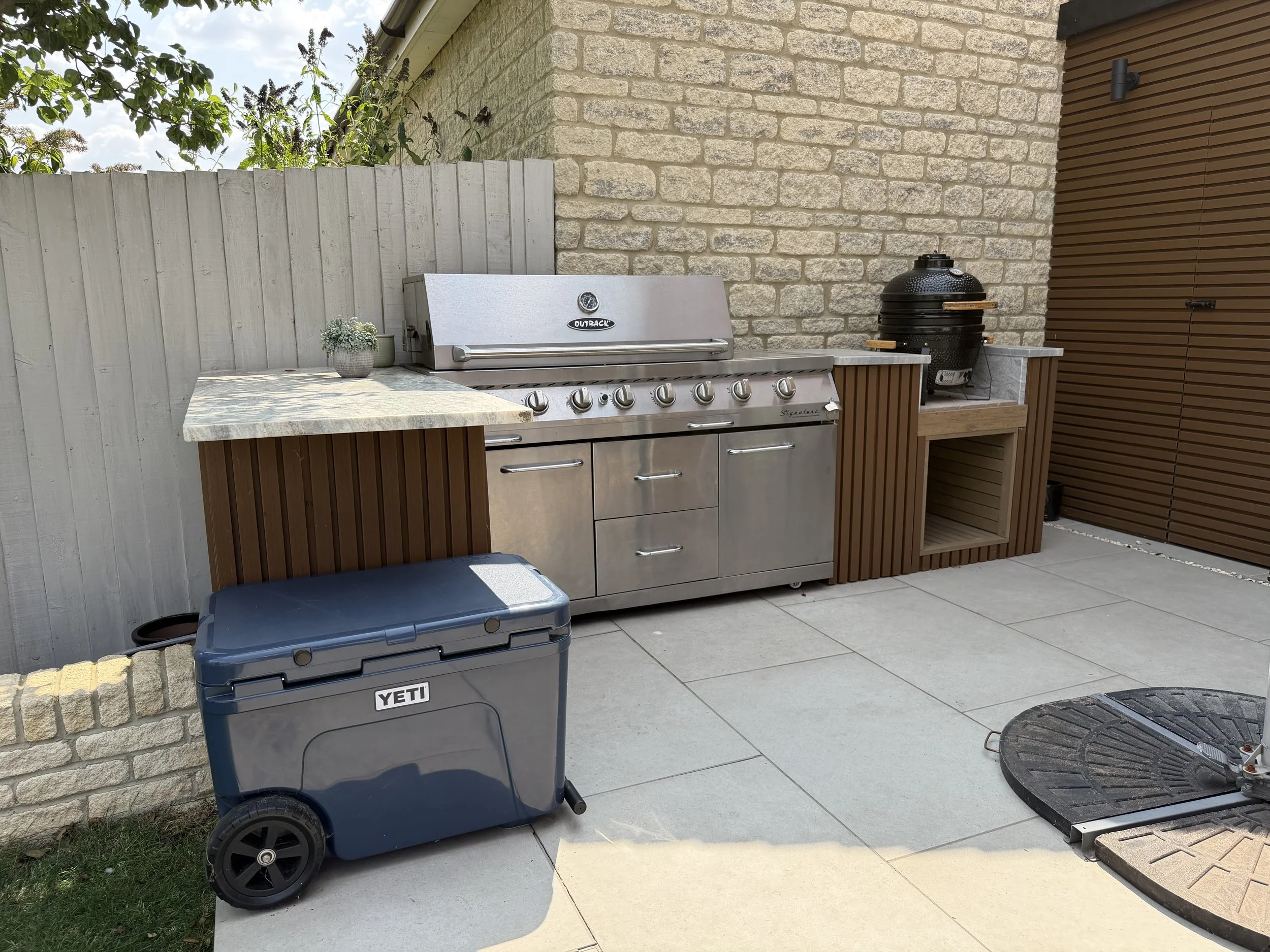 An outdoor patio area featuring a stainless steel outdoor grill with five control knobs and a thermometer on top, next to a sectional stone wall. To the right of the grill, there is a round black charcoal grill with a lid, situated on a wooden platform. A potted plant with small white flowers and a small green vase are on the marble countertop of the grill, which is attached to a wooden structure. In the foreground, a blue YETI trash or storage bin is on the concrete patio, with a black round table visible. A white fence and some leafy green trees are in the background.