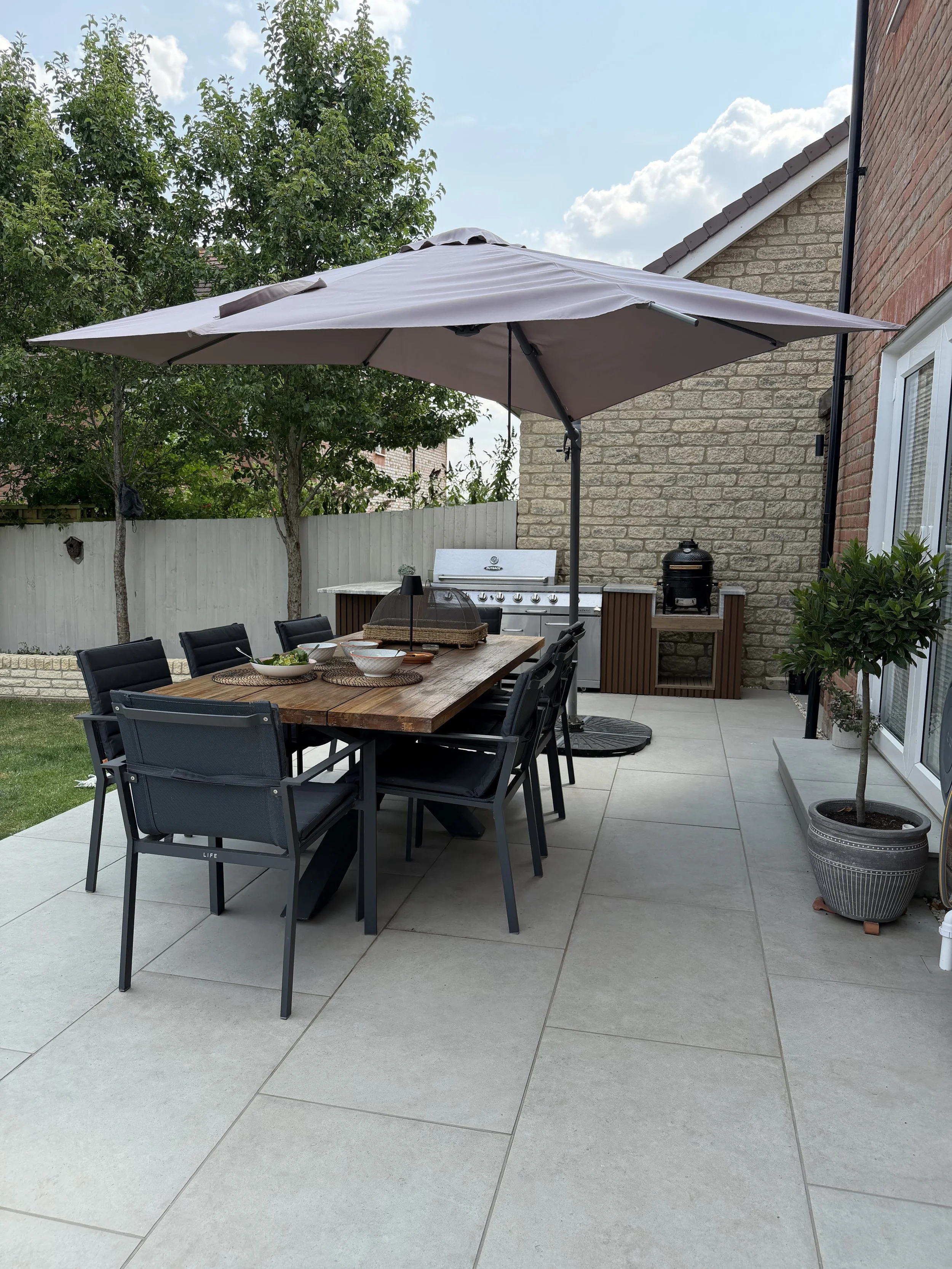 A backyard patio with a wooden dining table, black chairs, and a large umbrella providing shade. There are some bowls with food on the table. In the background, there is a grill, a small black smoker, a potted plant, and trees with a white fence, brick house, and cloudy sky.