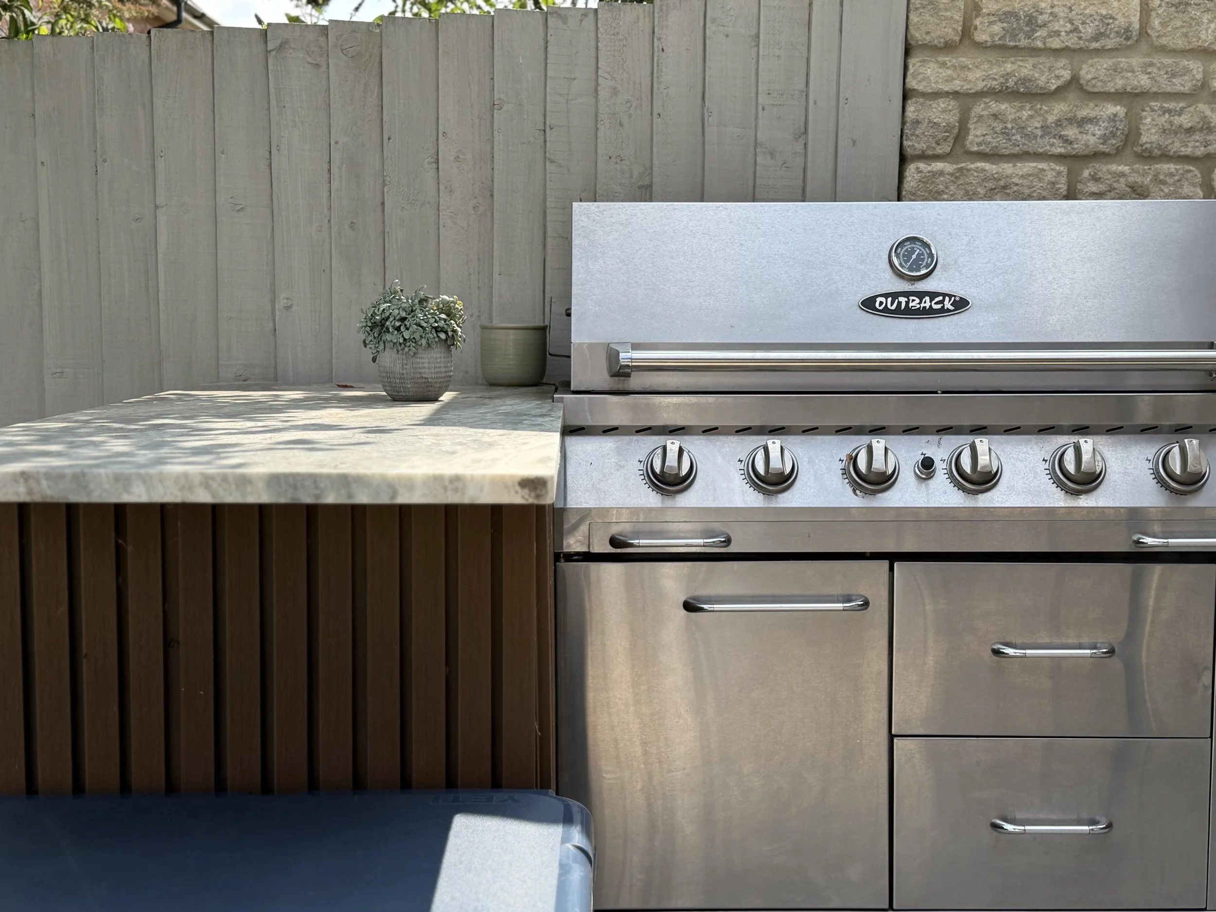 Outdoor kitchen with a stainless steel Outback barbecue grill, two potted plants on a stone countertop, and a wooden and stone fence in the background.