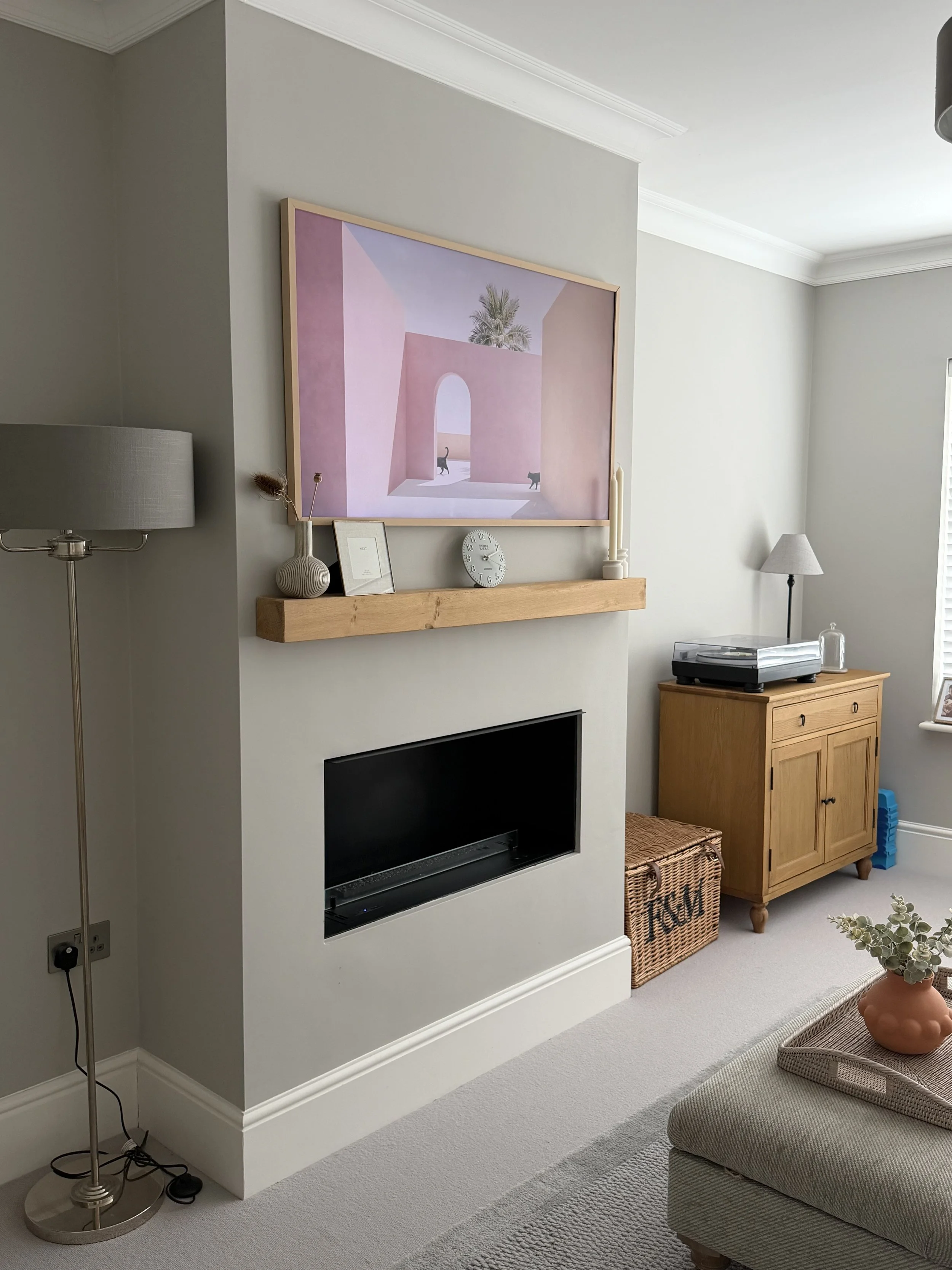 Living room with beige sofa, wooden cabinet, framed artwork of pink architecture and palm trees, white clock, candles, and drying rack, with a floor lamp and a television.