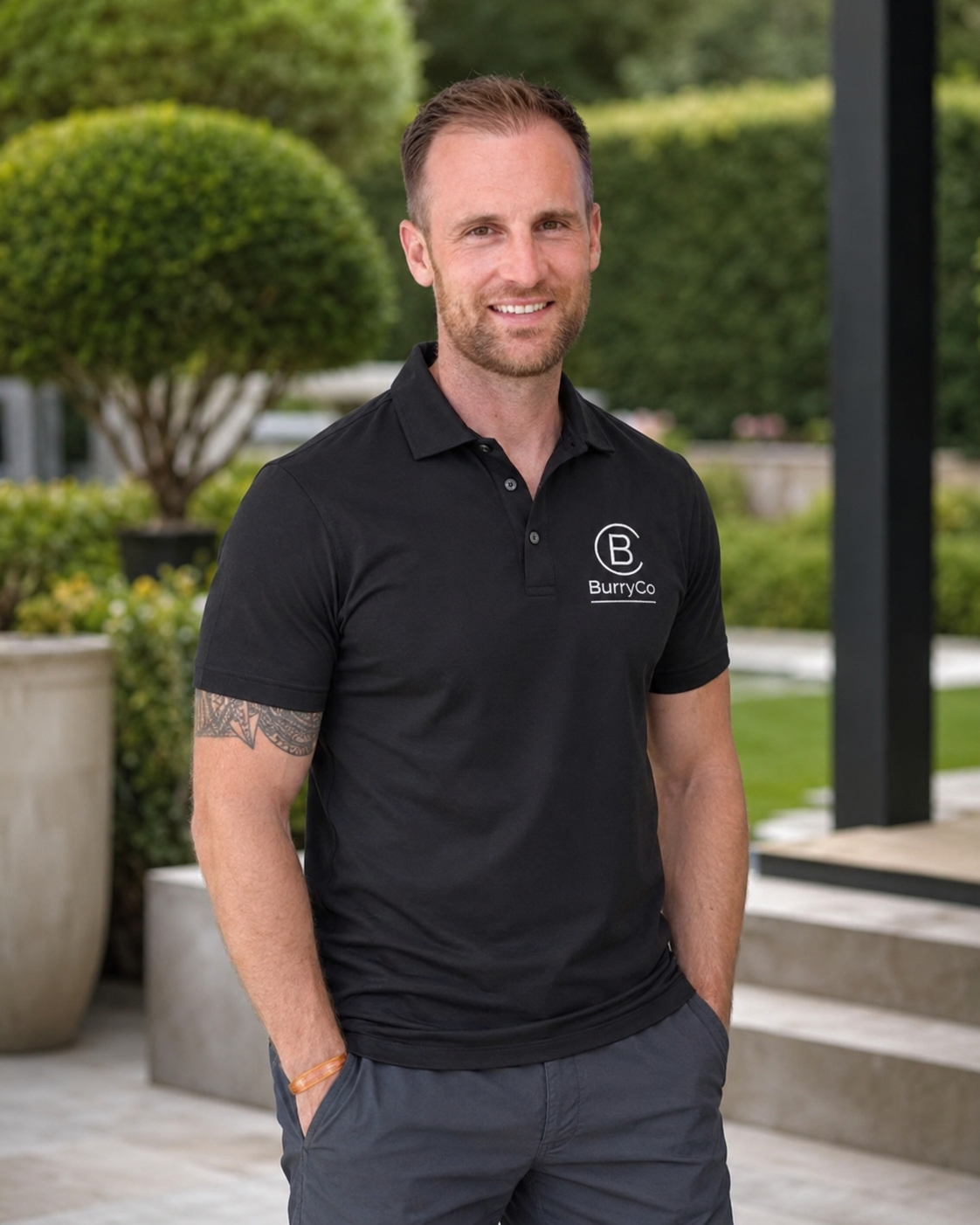 A smiling man with short brown hair and a beard, wearing a black polo shirt with the logo 'Burry Co' on it, standing outdoors near green bushes and trees in a landscaped area.