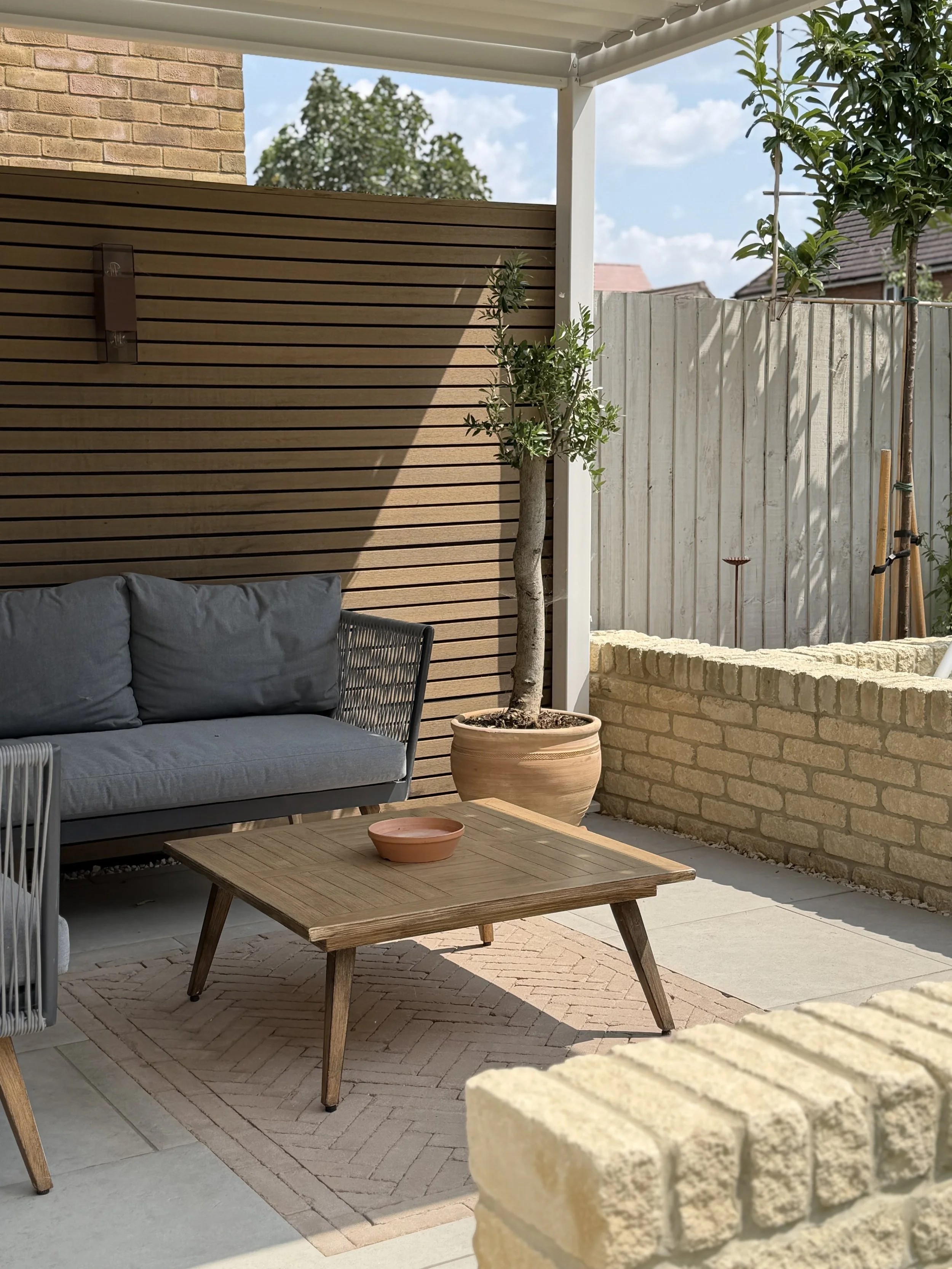 An outdoor patio area with a grey cushioned sofa, a wooden coffee table with an empty terracotta bowl, a potted tree, and a beige brick wall. There is also a wooden fence, some trees, and a partly cloudy sky visible in the background.