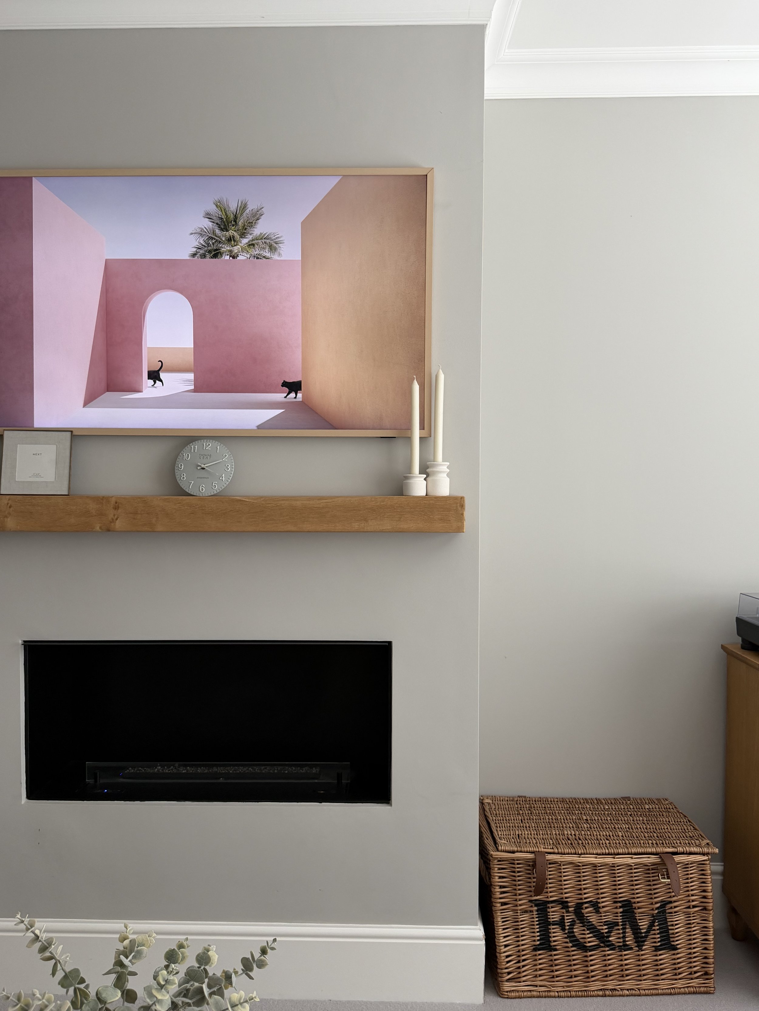 Living room wall with a large framed artwork of a pink architectural structure with an archway, a palm tree, and two black cats. Below the artwork is a wooden shelf with a small clock and a picture frame. To the right of the shelf, there are two white candles on a holder. To the right of the candles is a wicker storage basket with the word 'HOME' on it. There is also a black fireplace below the shelf and a small portion of a plant at the bottom of the image.