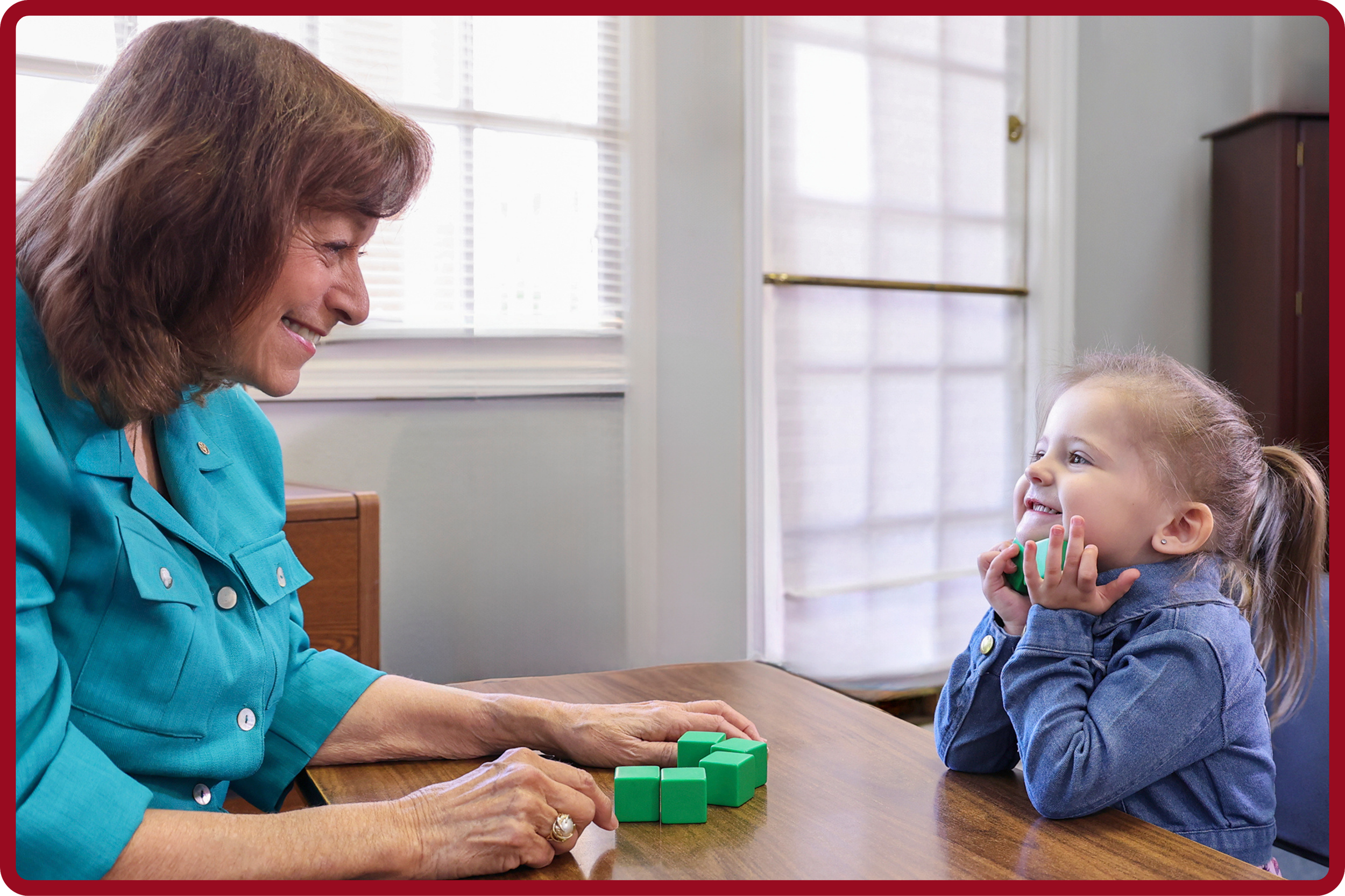 An older woman and a young girl playing a game with green blocks at a wooden table indoors, with bright natural light coming through windows in the background.