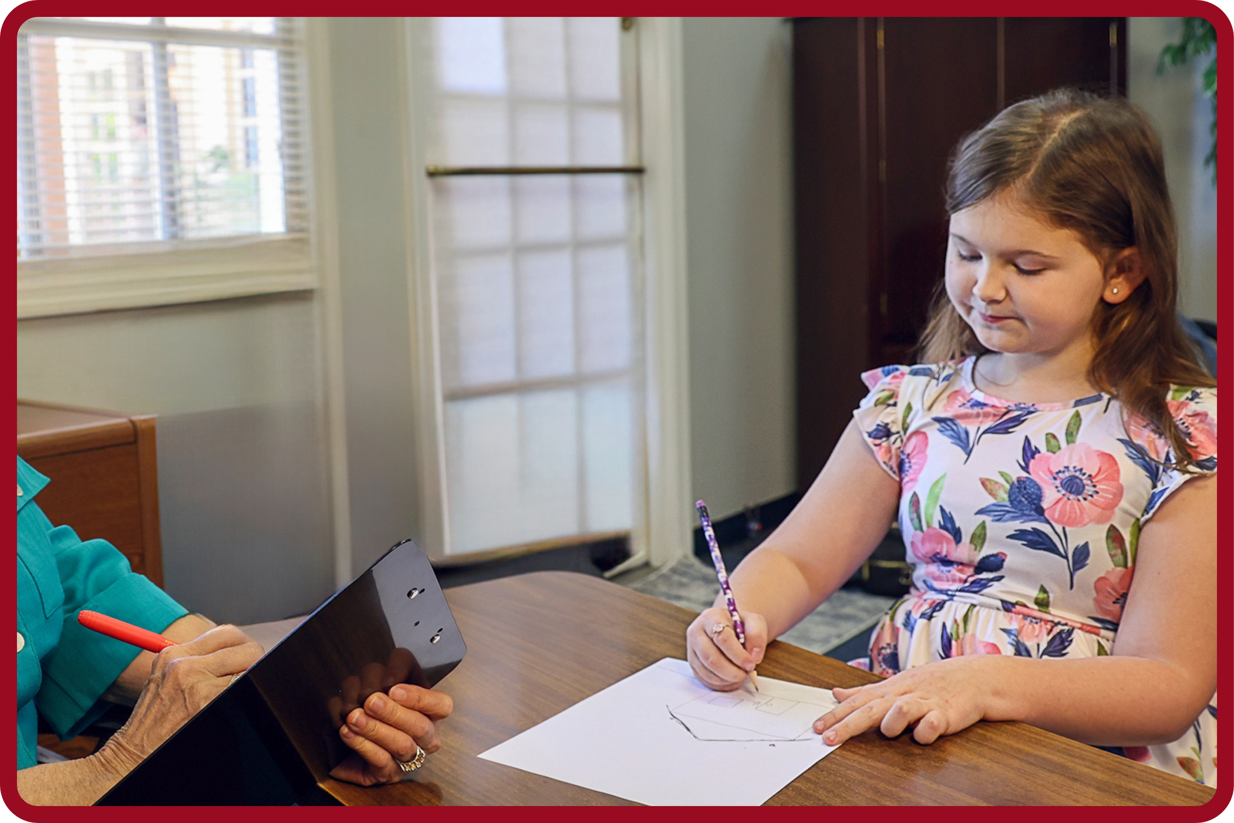 Young girl with brown hair and earrings wearing a floral dress, sitting at a table, drawing on a paper with a pencil, while an adult holds a tablet in front of her.