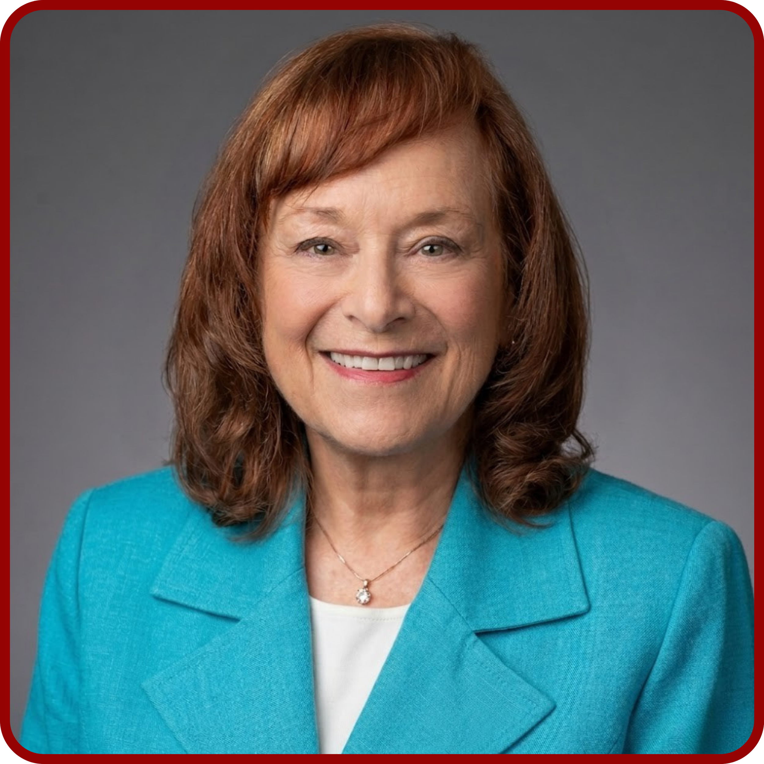 A woman with reddish-brown hair smiling, wearing a teal blazer, white top, and a necklace with a round pendant on a gray background.