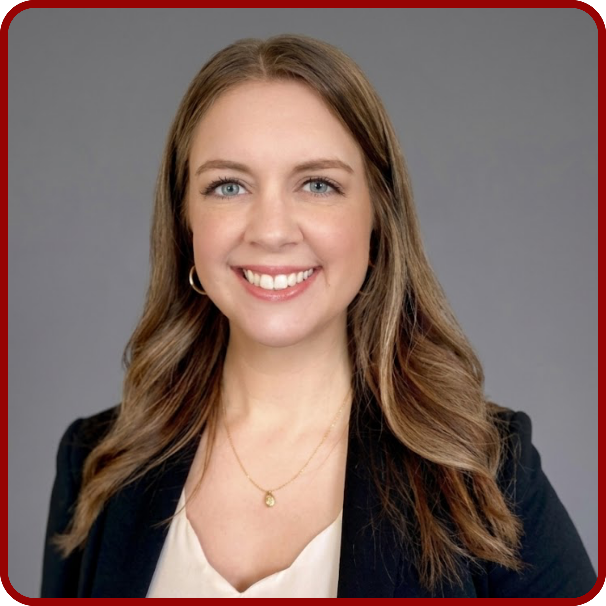 Close-up of a woman with long, wavy brown hair, blue eyes, light makeup, wearing a black blazer, white blouse, gold necklace, and gold hoop earrings, smiling in front of a gray background.