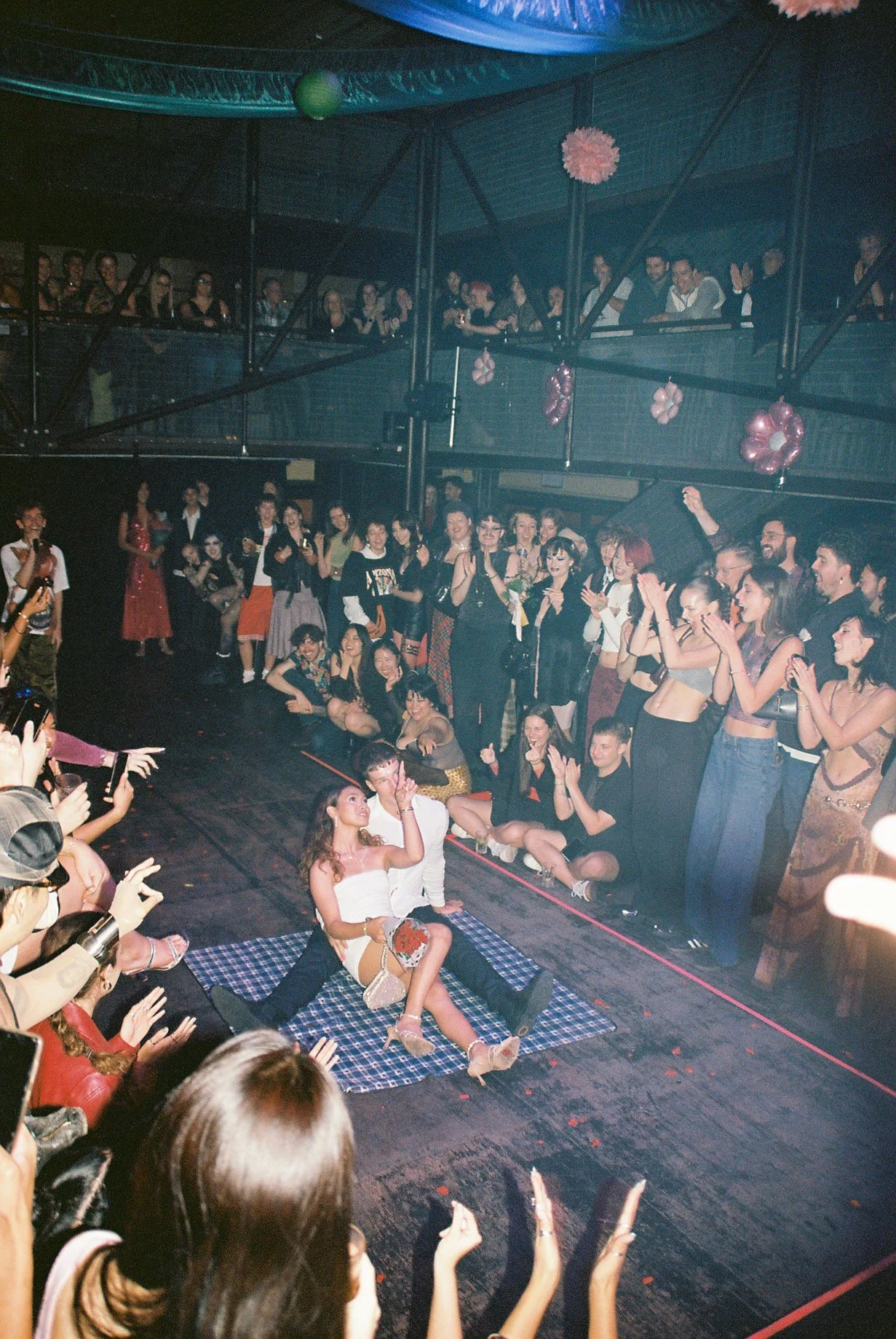 A lively party scene with a large group of people, some standing on a balcony and others sitting or kneeling on the floor, clapping and smiling, with a couple sitting on a checkered blanket in the foreground.