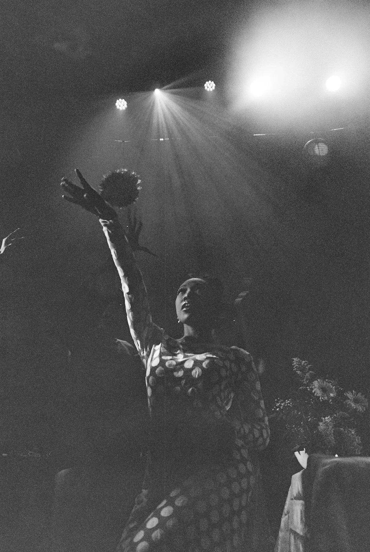 A person with short hair and a polka-dot dress, raising an arm with a peace sign gesture, standing in front of a stage with bright stage lights.