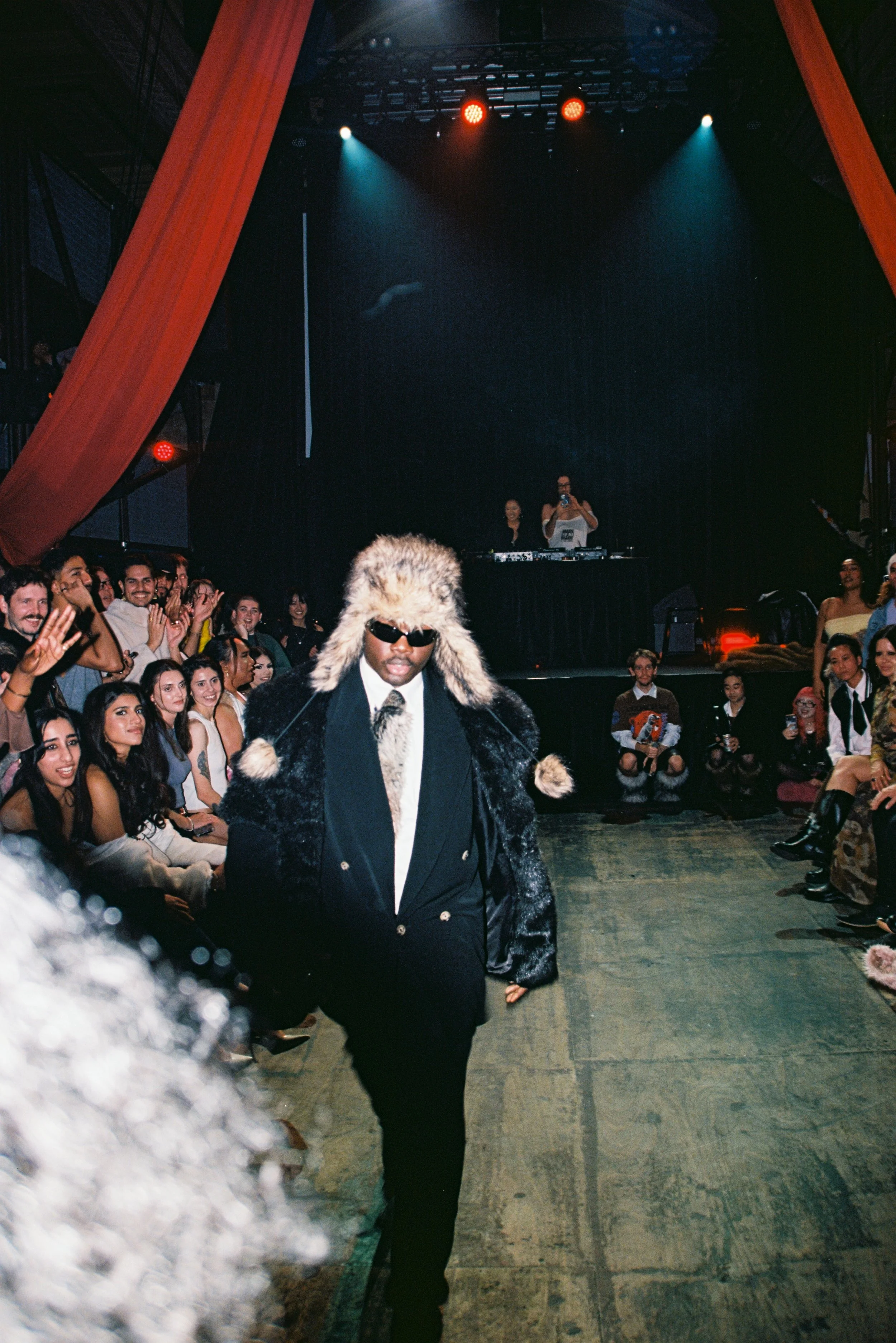 A man wearing sunglasses, a fur hat, and a coat walking through a crowded venue with dance floor and stage in the background.