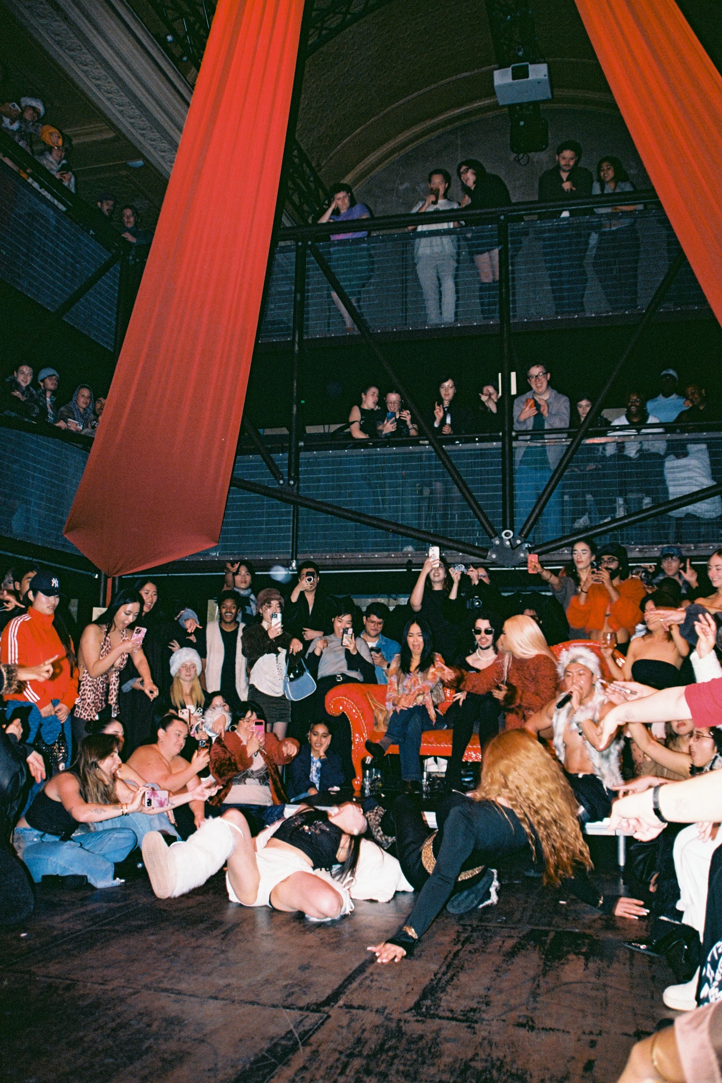 A lively indoor party scene with a large crowd of people, some seated and some standing, many taking photos or videos, with a performer on the floor in front of the audience. The venue has an industrial style with balconies and large orange fabric decorations.