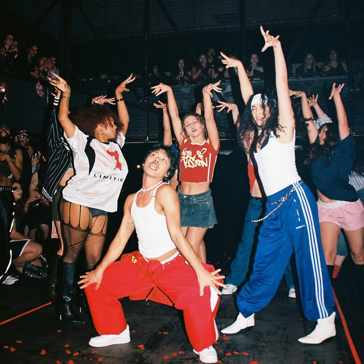 Group of women dancing at a party or club, some with arms raised, vibrant atmosphere, multiple levels with people watching from above.